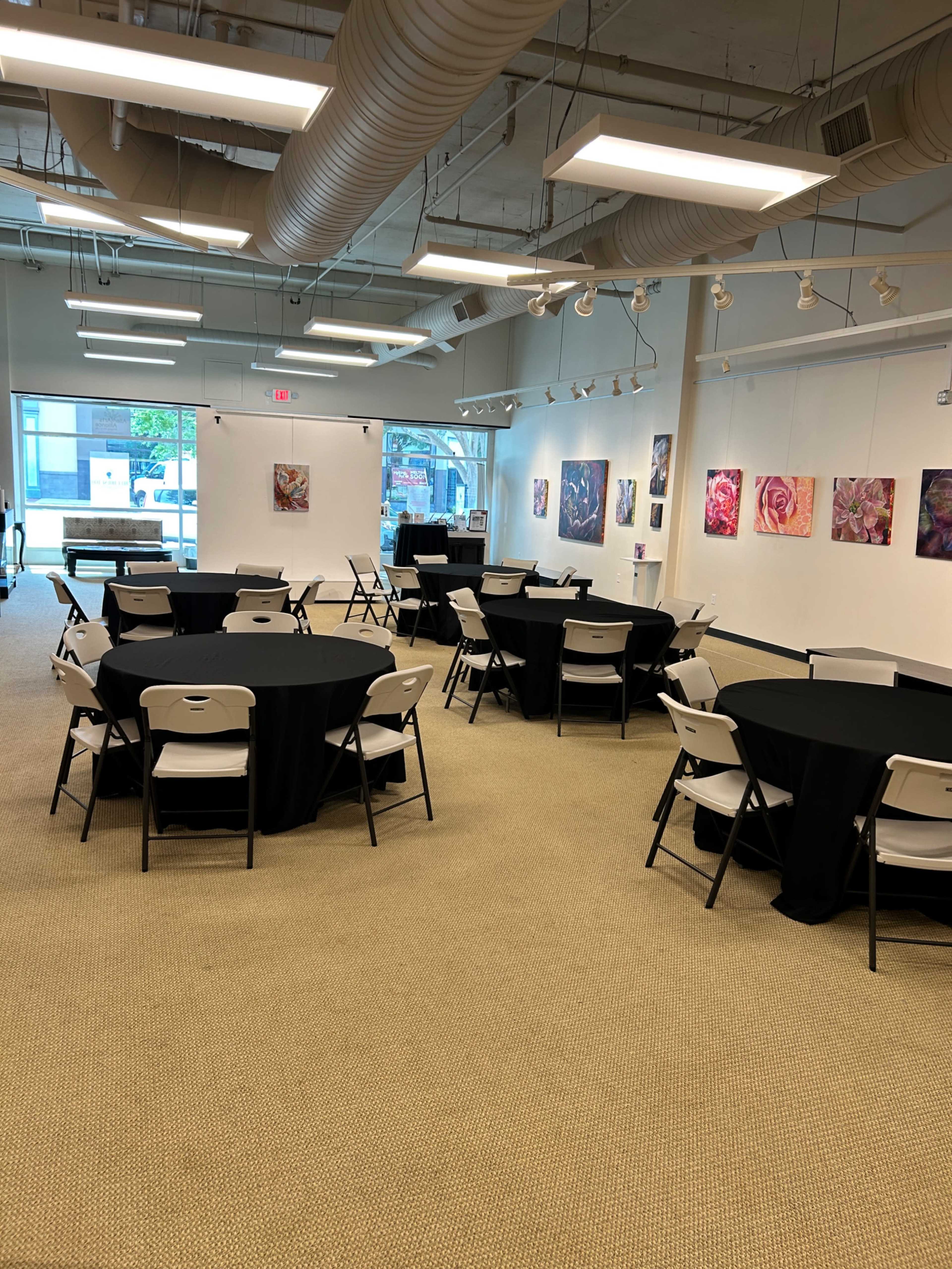 The image shows a spacious room with several round tables covered with black tablecloths and a few pieces of artwork hung on the walls.