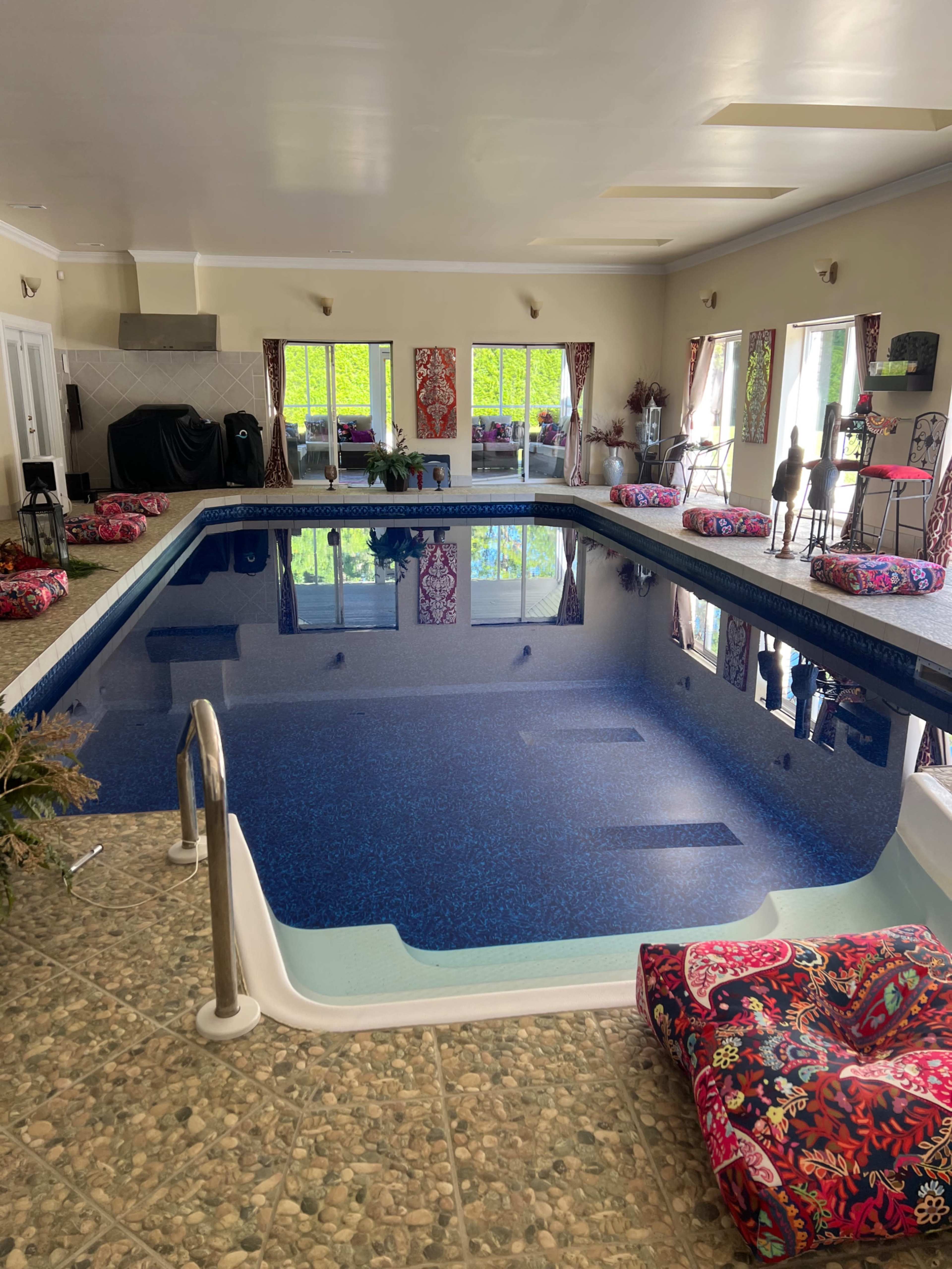An indoor swimming pool surrounded by colorful cushions and large windows with green views.