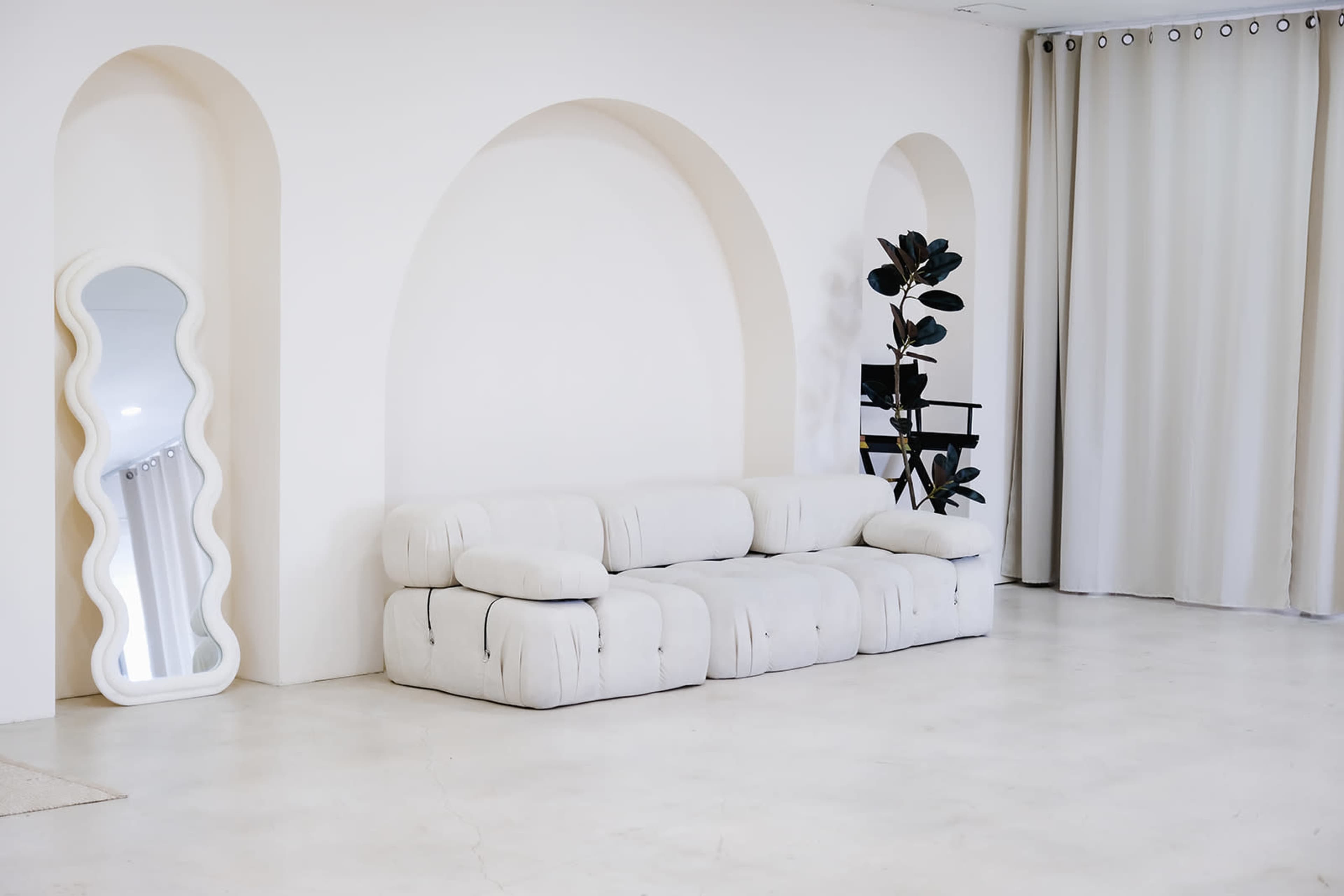 The image shows a minimalist interior featuring a white sofa, a tall potted plant, a decorative mirror, and beige curtains against a light-colored wall with archways.