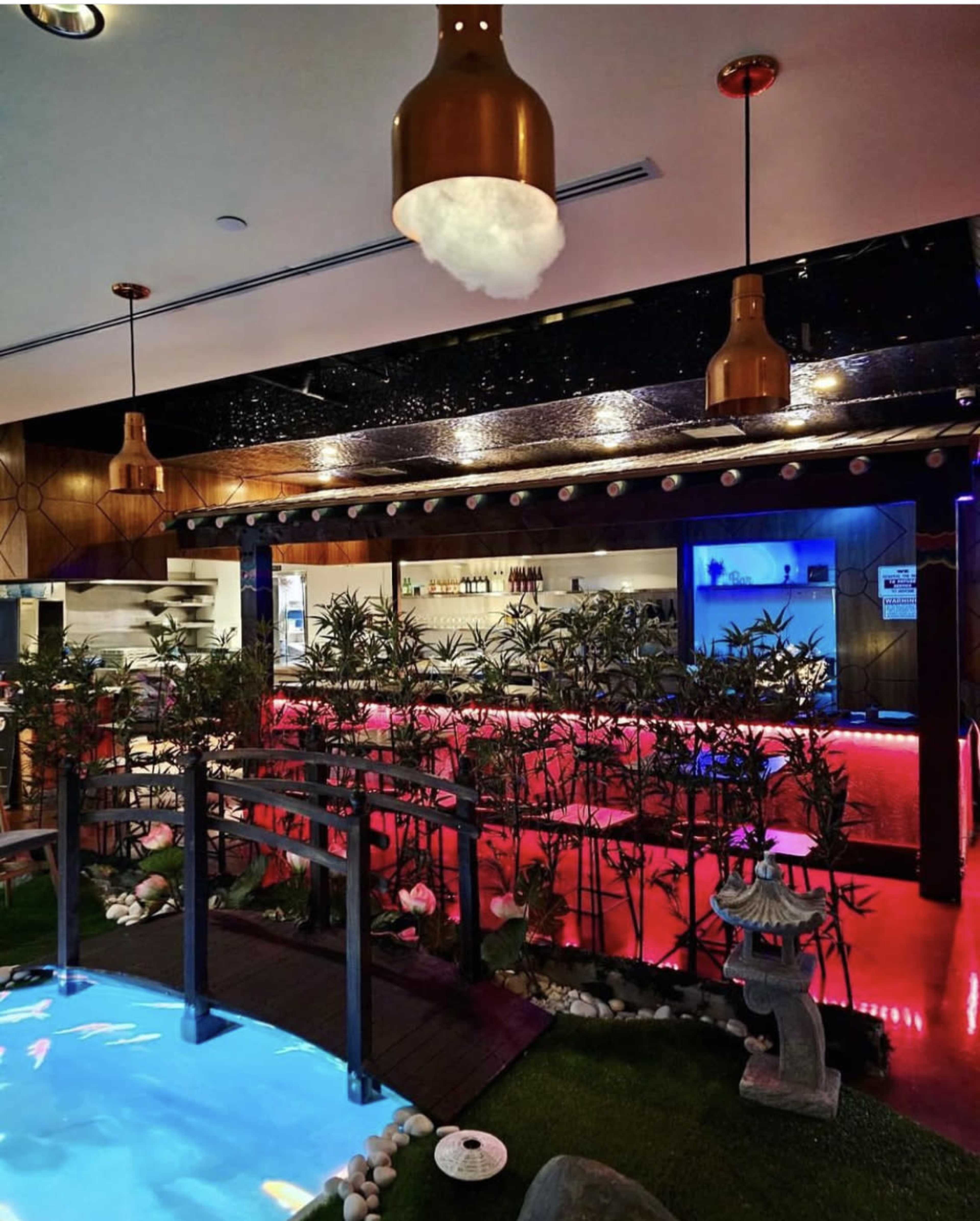A modern interior space featuring a small wooden bridge over a blue pond, surrounded by bamboo plants and illuminated by decorative pendant lights.