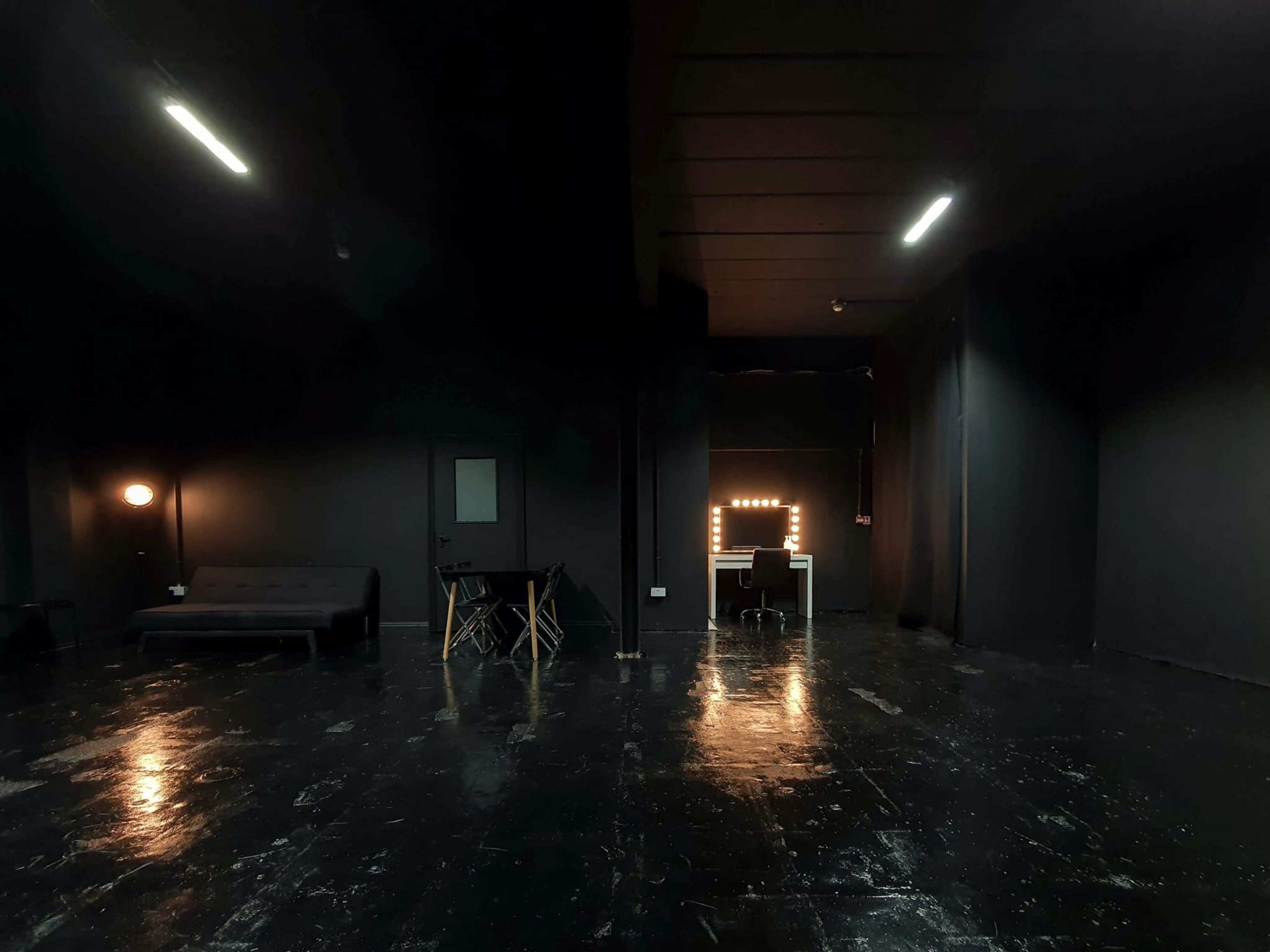 A dimly lit room with dark walls, a sofa on the left, a table and chairs in the center, and a brightly lit vanity mirror against the back wall.