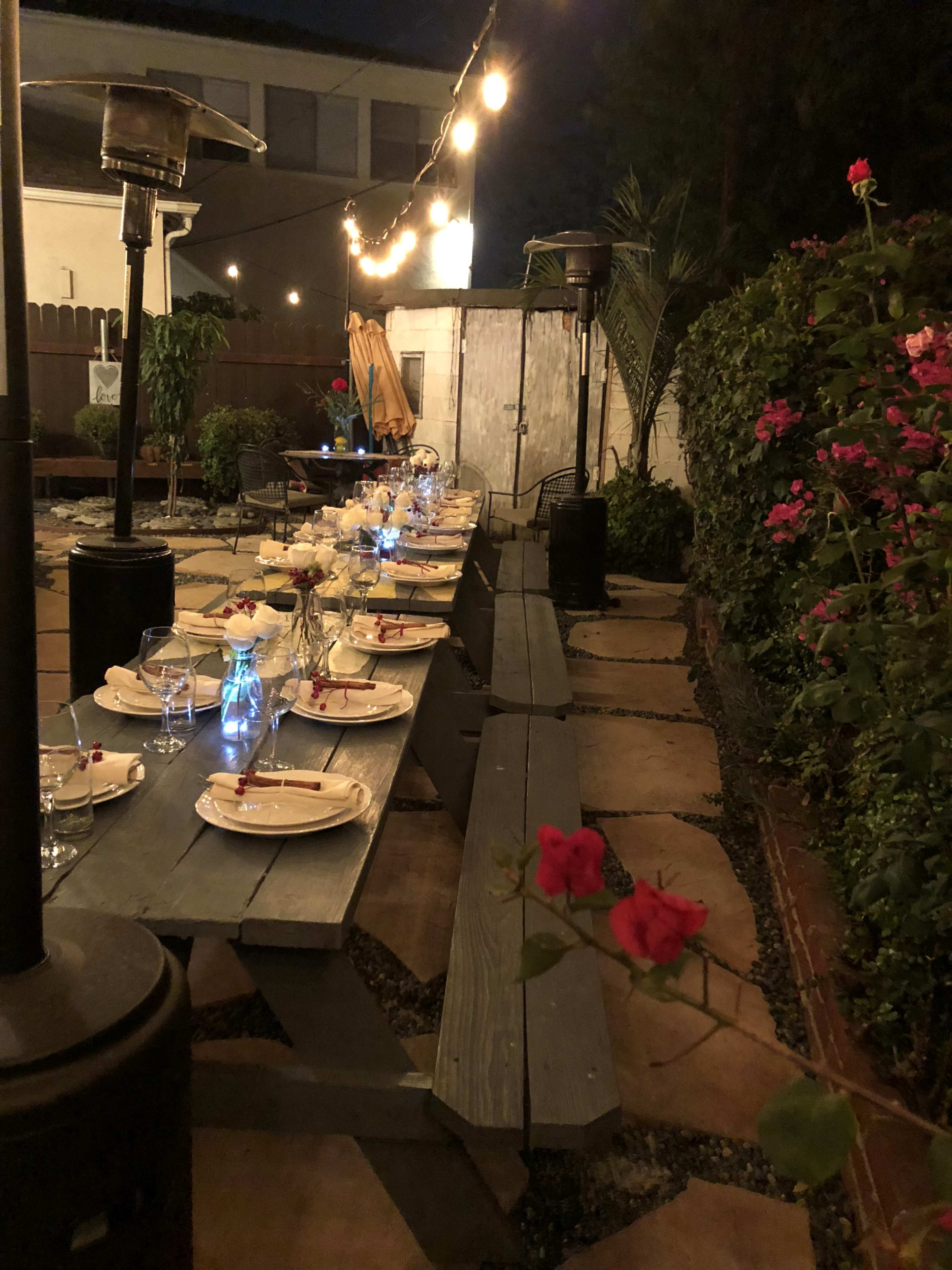 A long outdoor dining table is set with plates and glasses under string lights, surrounded by plants and flowers in a dimly lit garden.