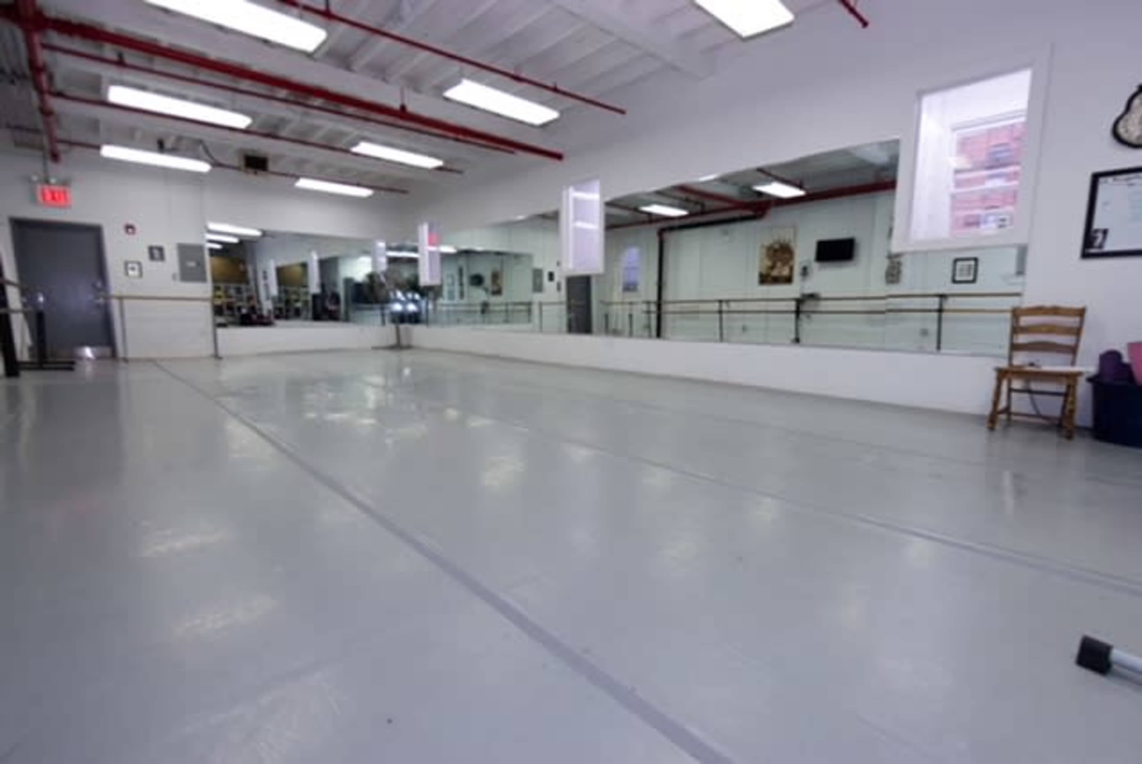 The image depicts an empty dance studio with gray flooring and mirrored walls, illuminated by natural light from large windows.