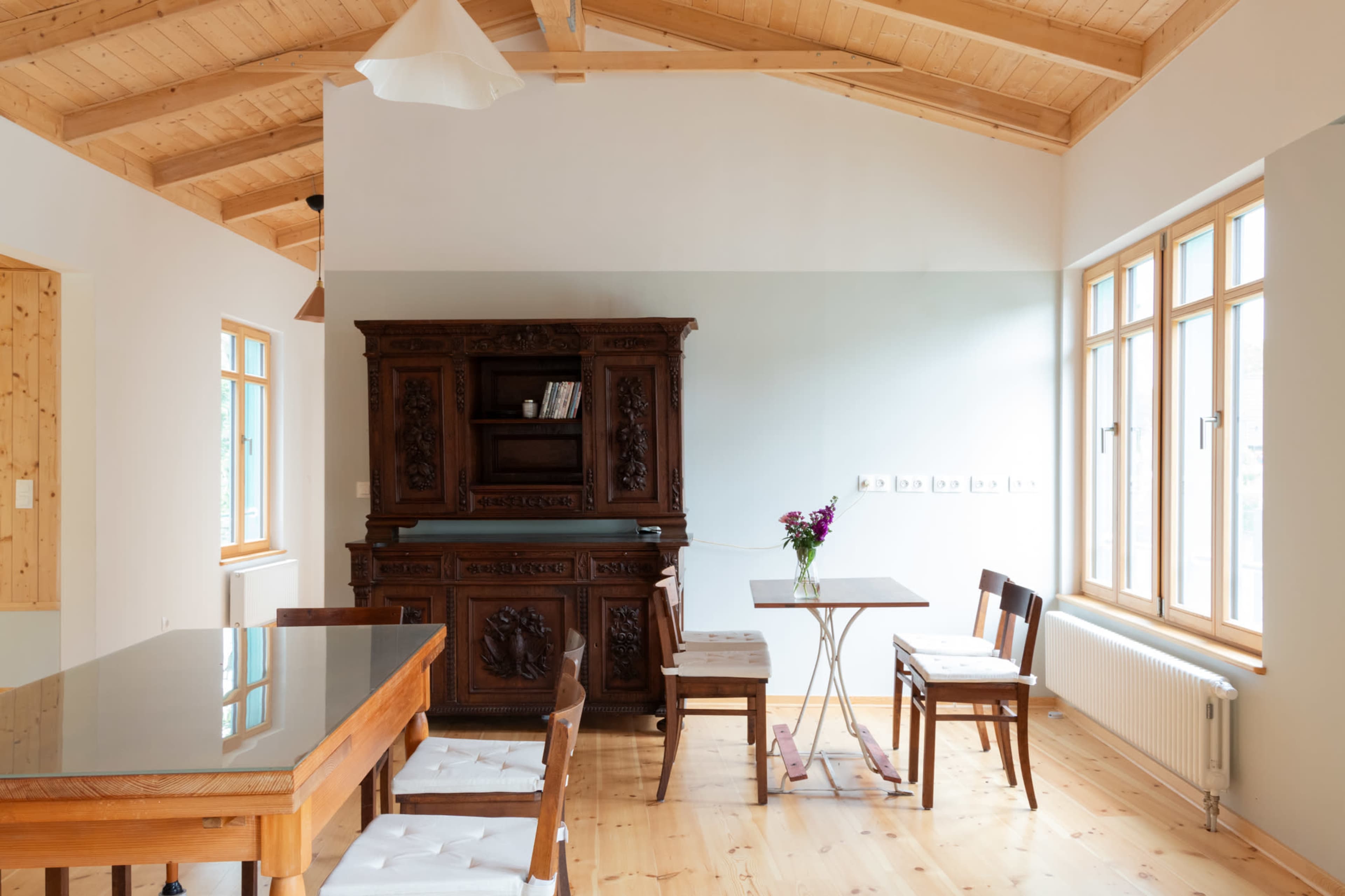 The image shows a bright, minimalist dining area featuring a wooden table, a vintage cabinet, and several wooden chairs under a sloped wooden ceiling.