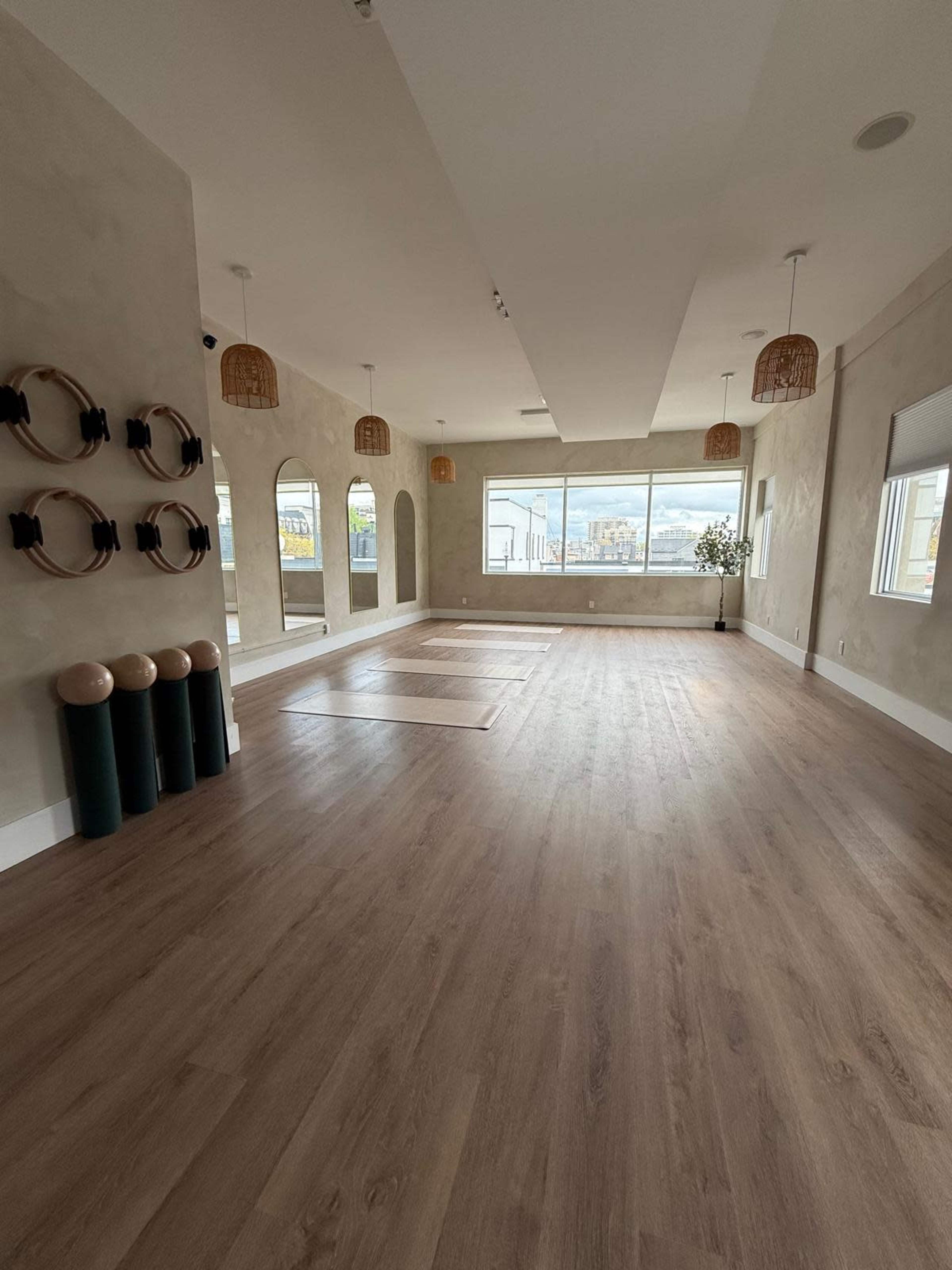 The image shows a spacious indoor studio with light wooden flooring, large windows, and decorative elements like rattan light fixtures and wall mirrors.