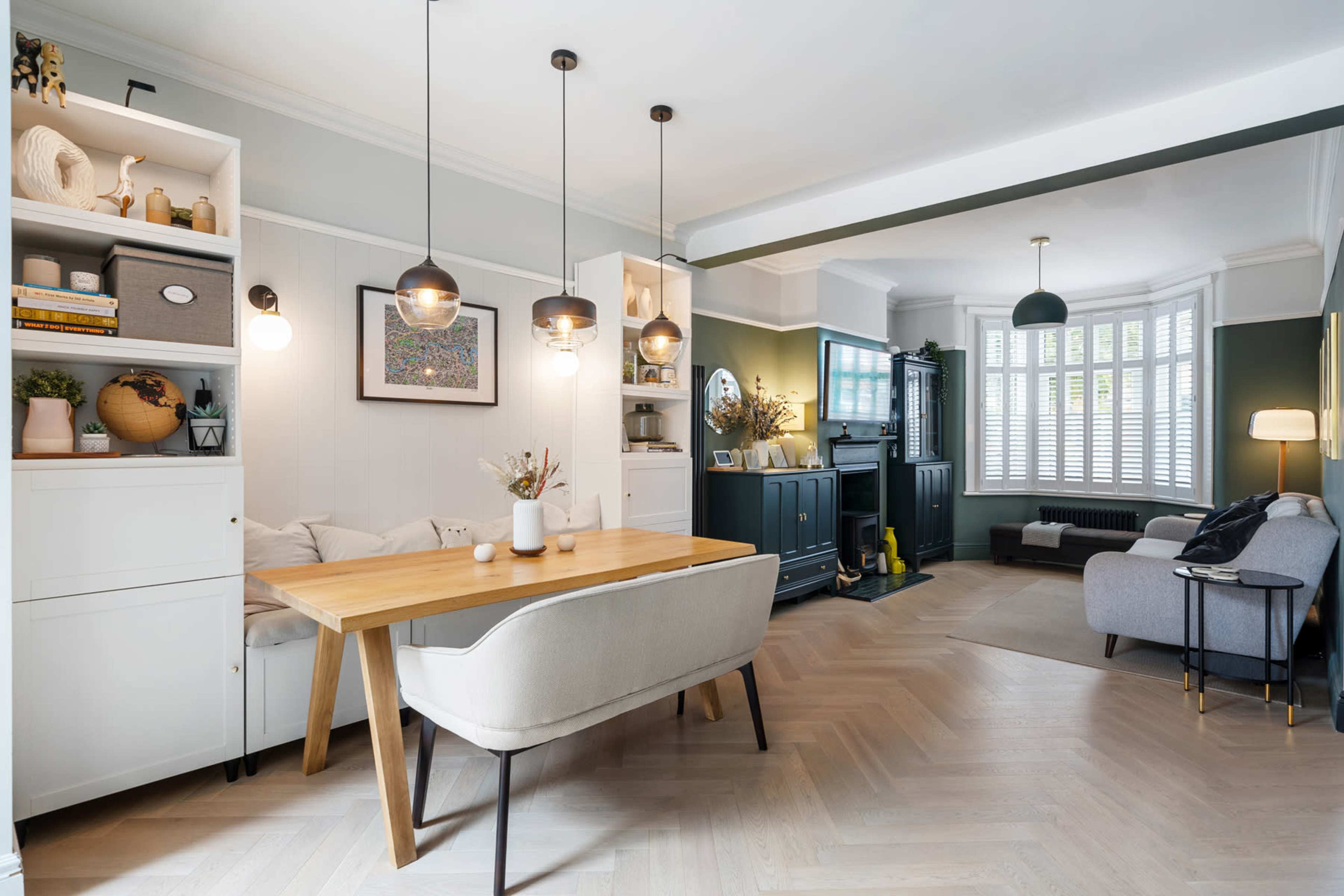 A modern living room and dining area features a light wood dining table, shelving units, and a gray sofa, with large windows allowing natural light.