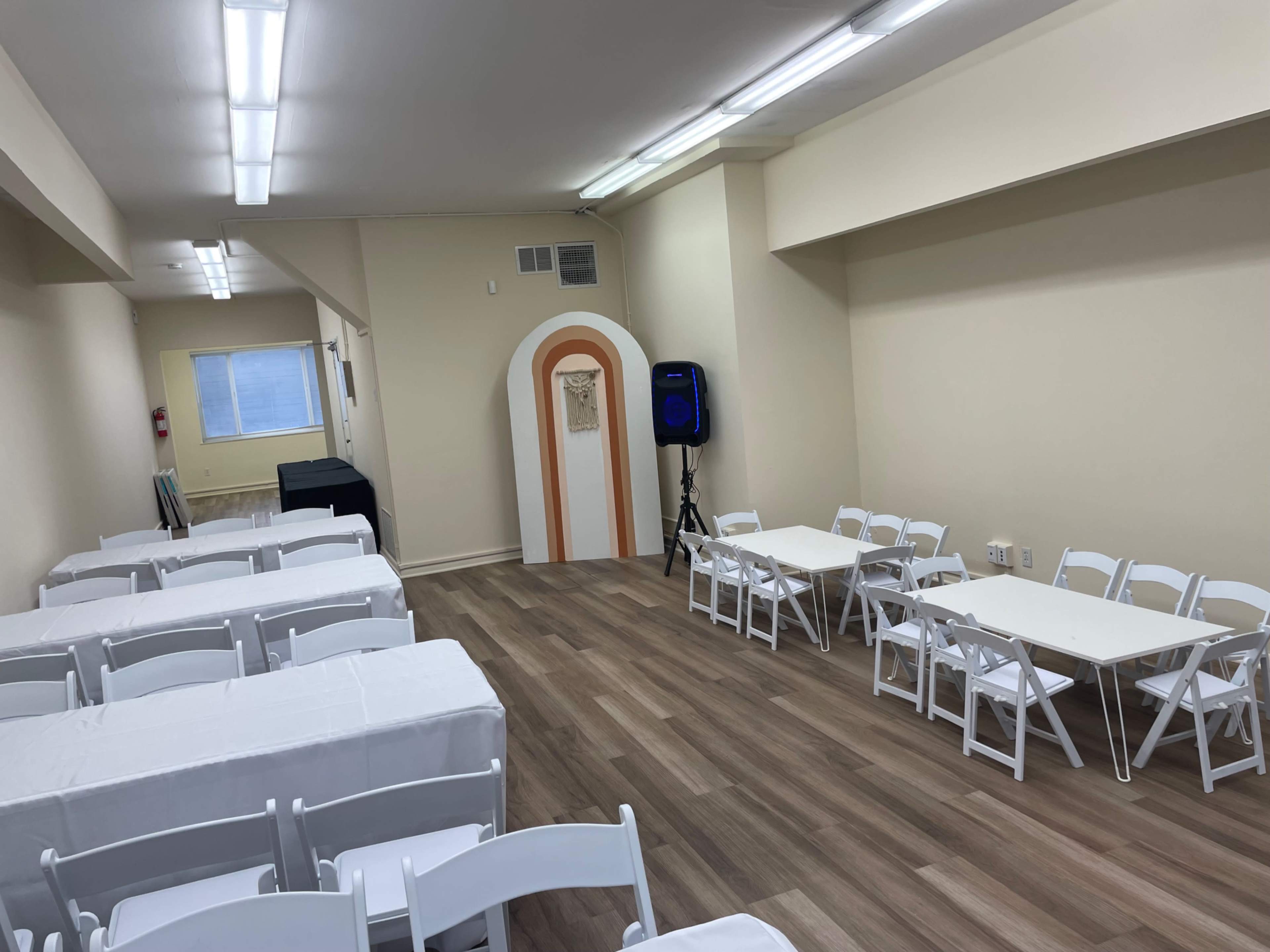 A spacious room with white folding chairs and tables arranged on a wooden floor, illuminated by fluorescent lights.