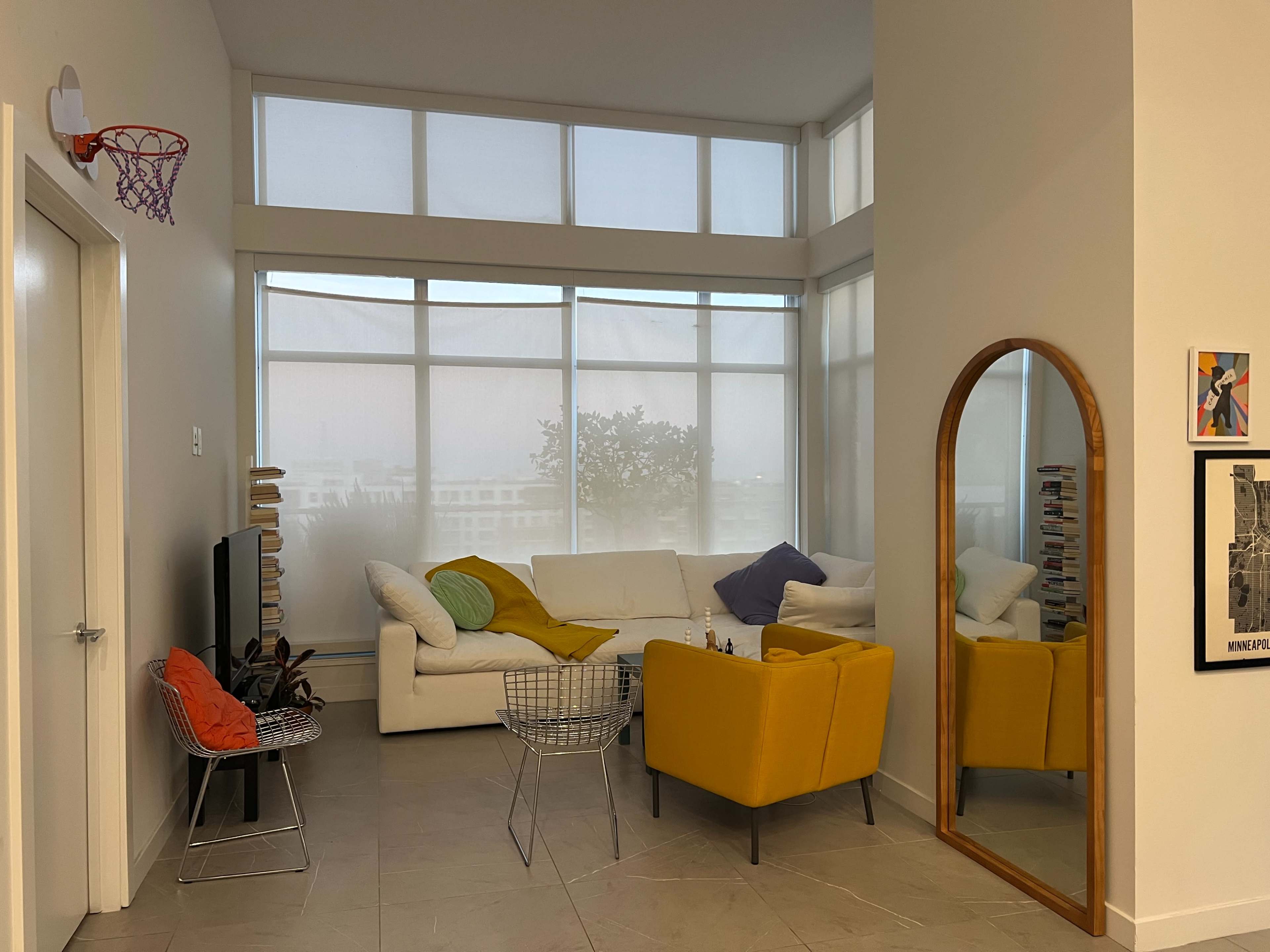 A modern living room features a white sectional sofa, a yellow armchair, a wall-mounted basketball hoop, and large windows that provide natural light.