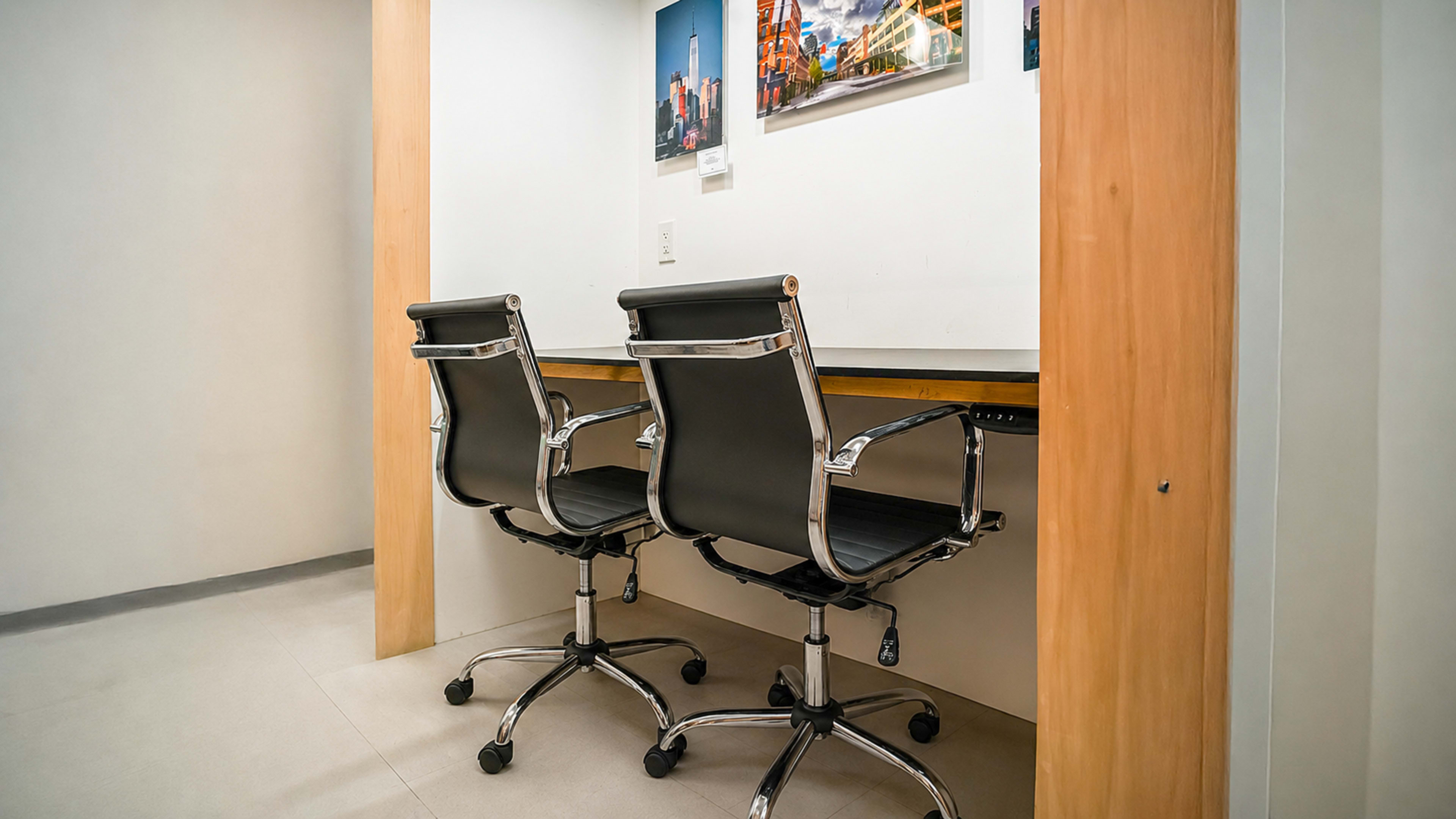 The image shows a minimalist workspace with two black office chairs positioned in front of a wooden desk against a light-colored wall.