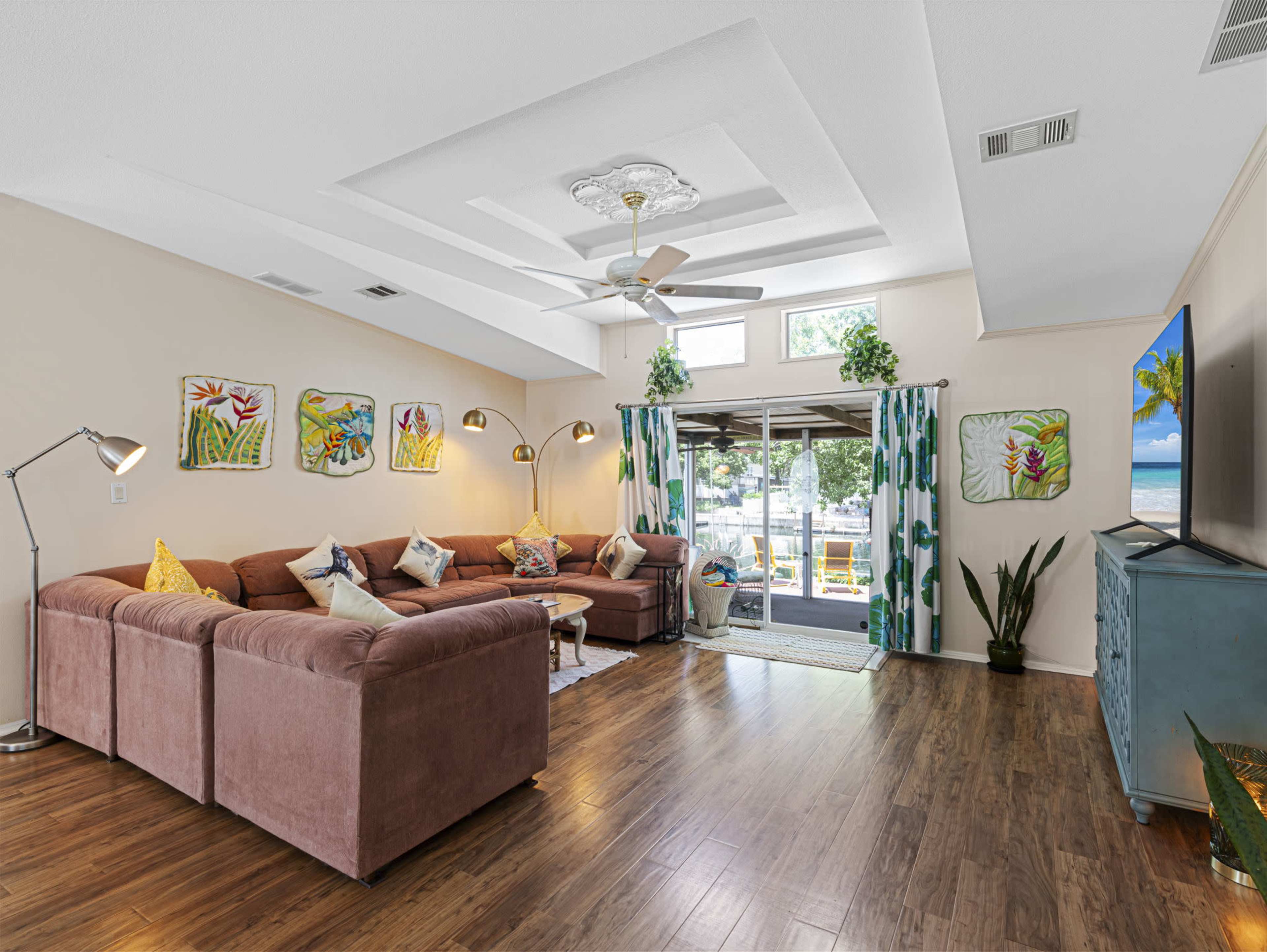 A brightly lit living room features two brown sofas, decorative artwork on the walls, and large windows with green curtains.