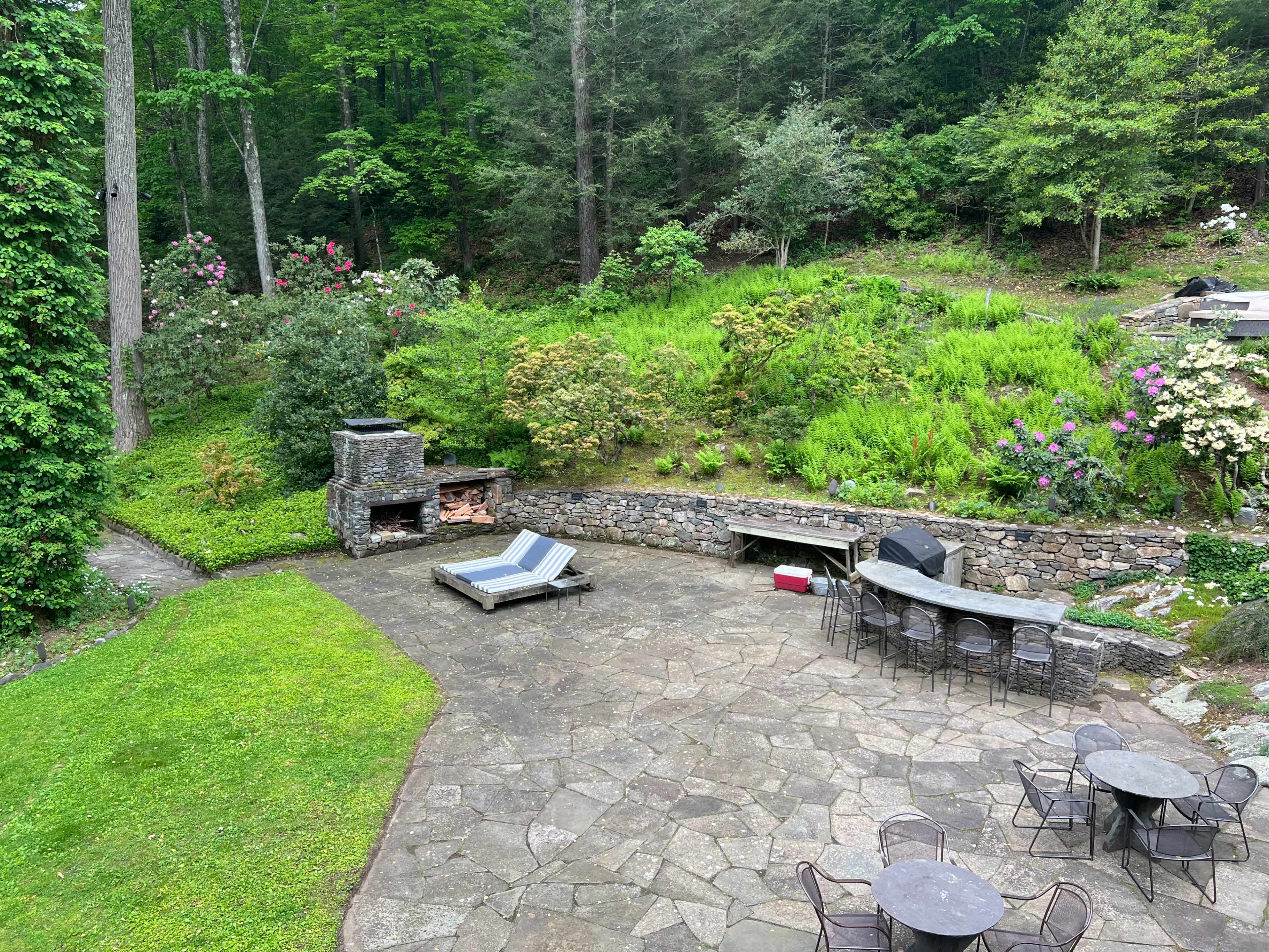 A stone patio area surrounded by greenery, featuring lounge chairs, an outdoor bar with stools, and a stone fireplace.