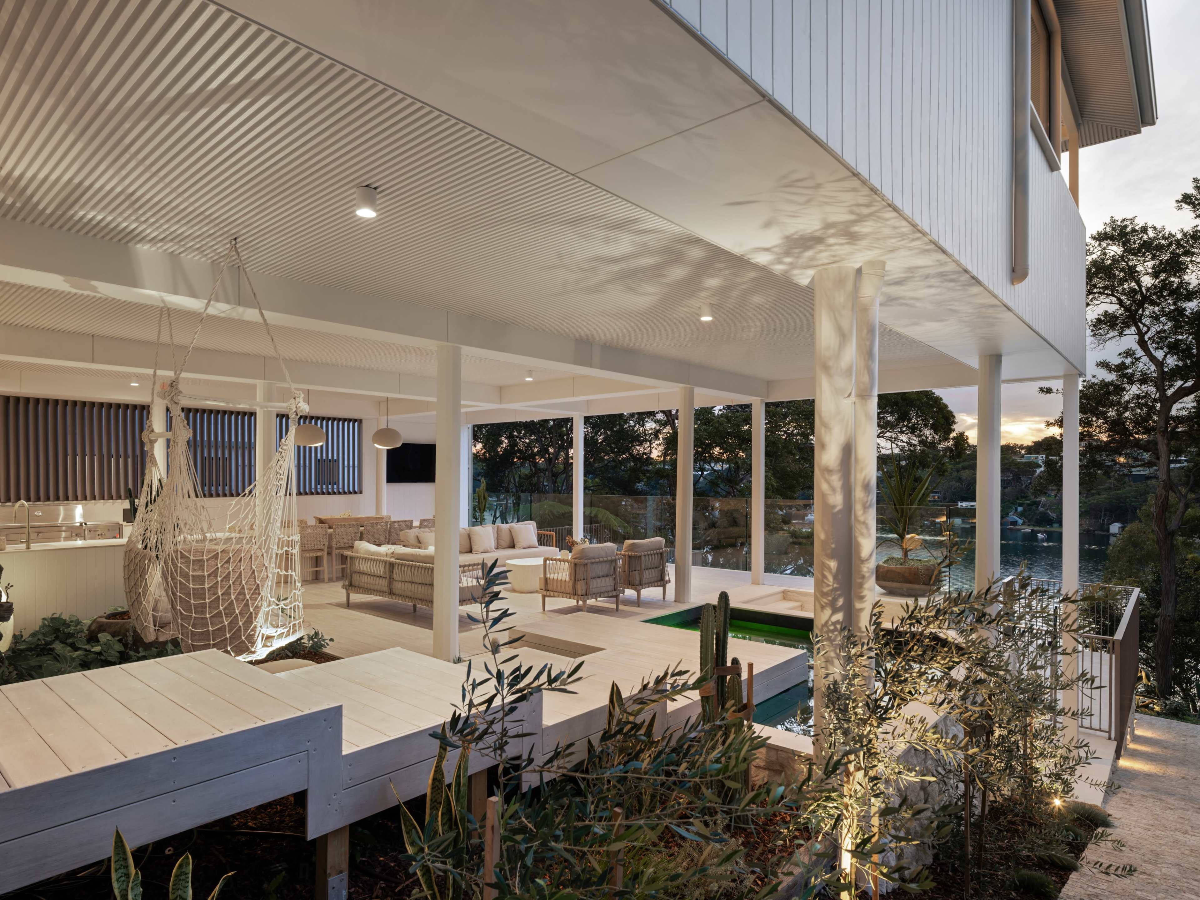 The image shows a modern house with a deck and seating area, surrounded by greenery and featuring a small pool.