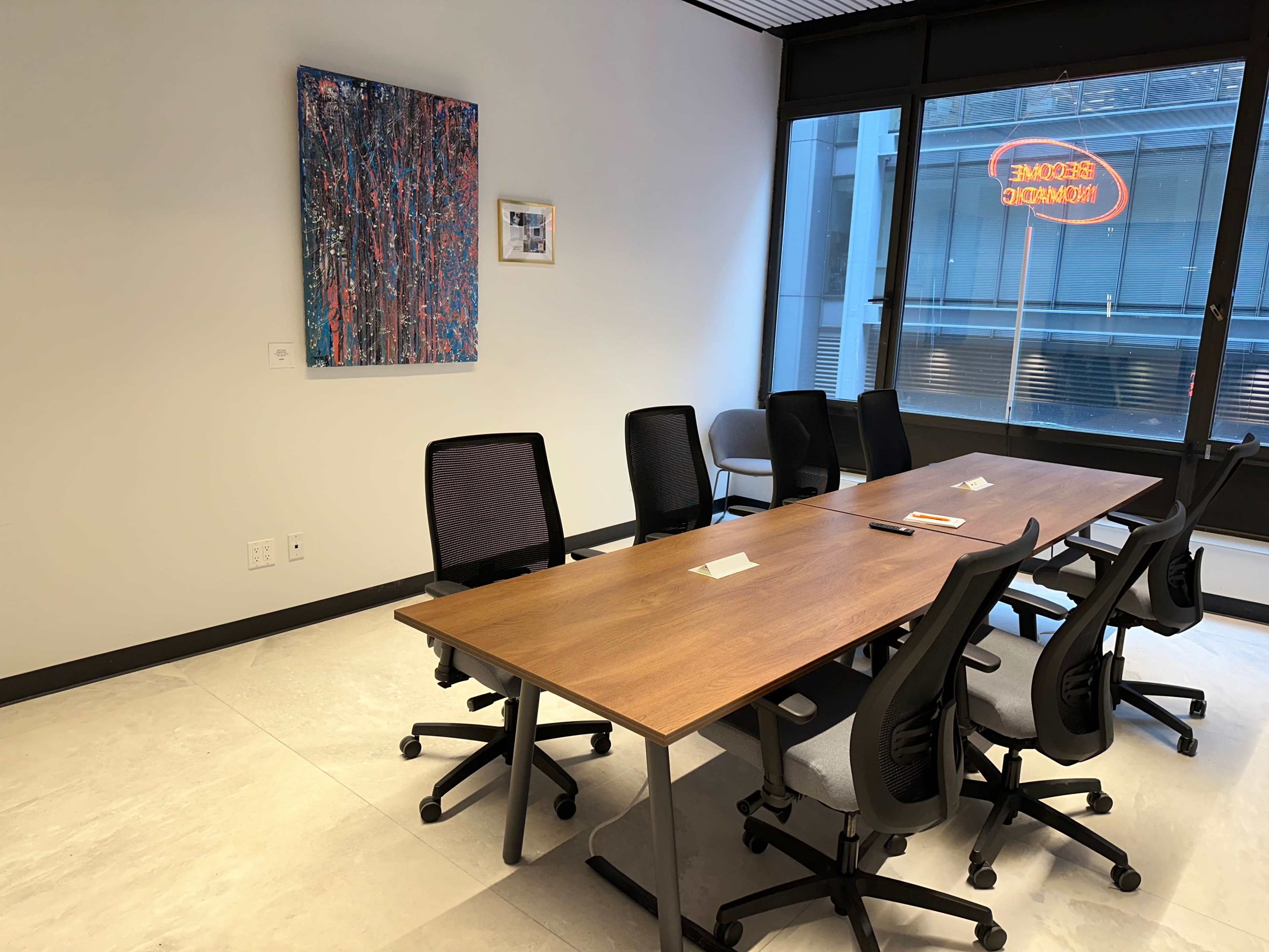 A conference room features a long wooden table surrounded by six office chairs, with two pieces of artwork on the wall and a neon sign visible through the window.