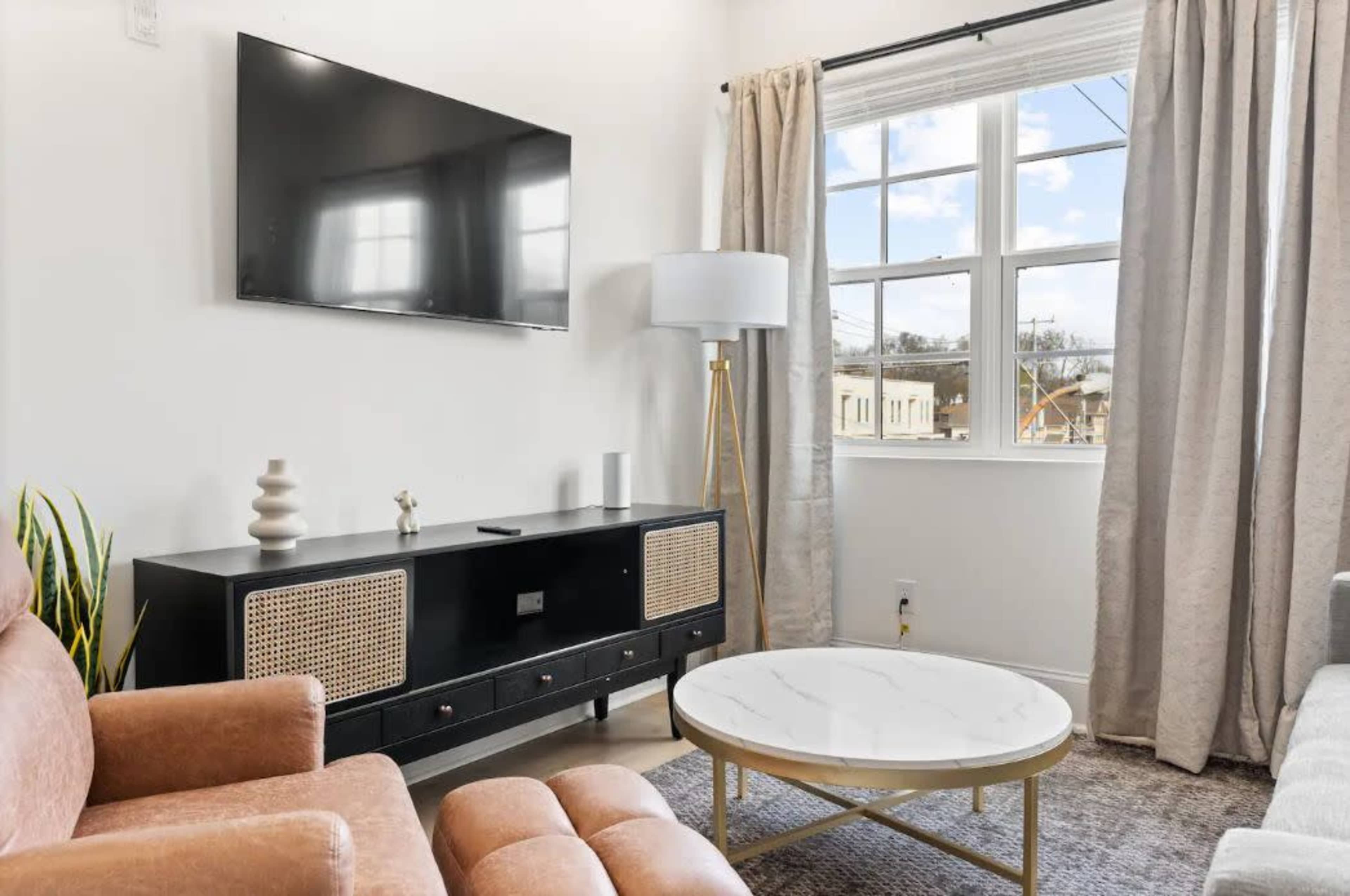 A modern living room features a wall-mounted television, a sleek coffee table, a comfortable armchair, and large windows with natural light filtering through.