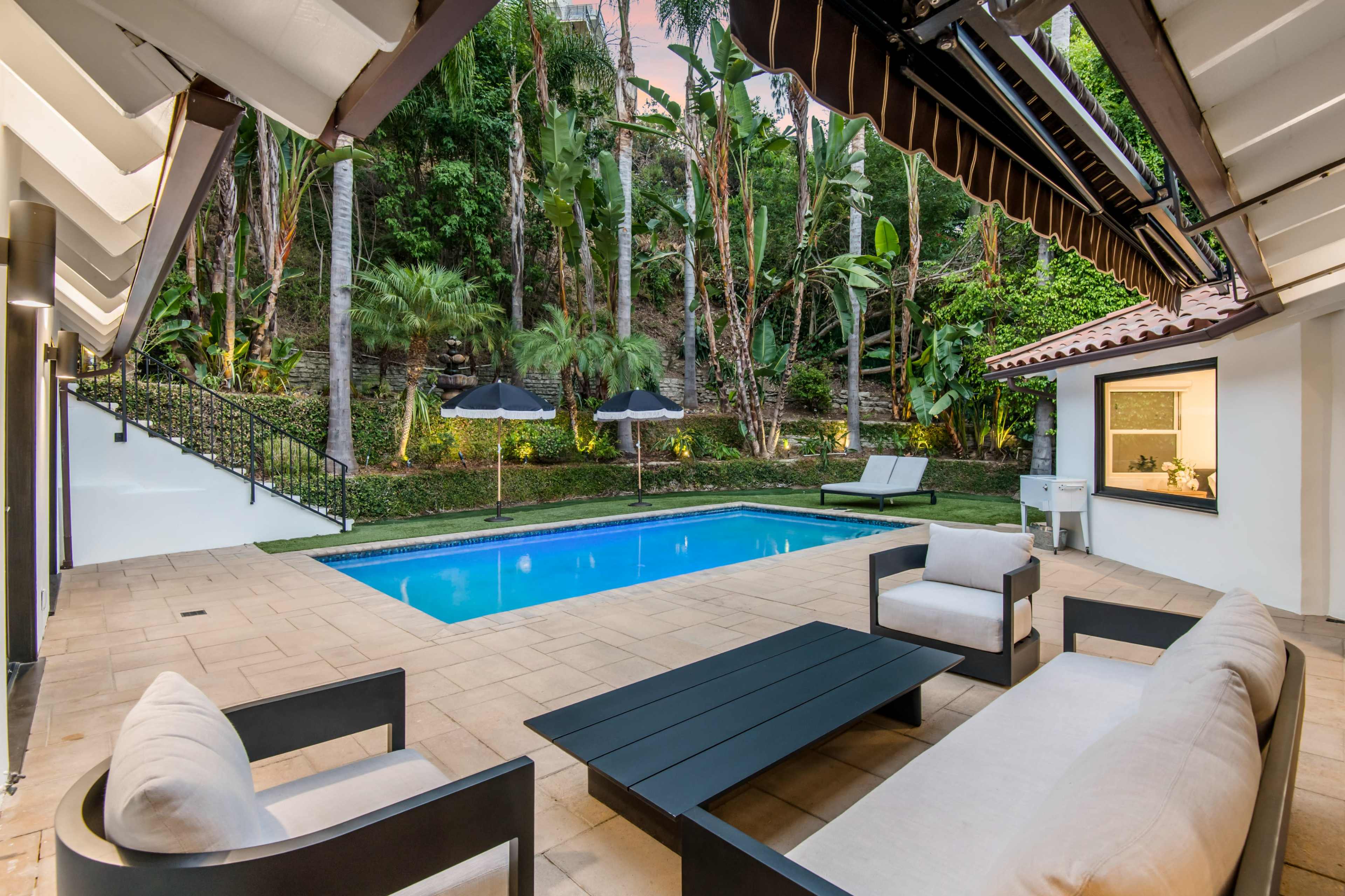 A modern patio features a pool surrounded by tropical plants, with seating areas and umbrellas in a serene outdoor setting.