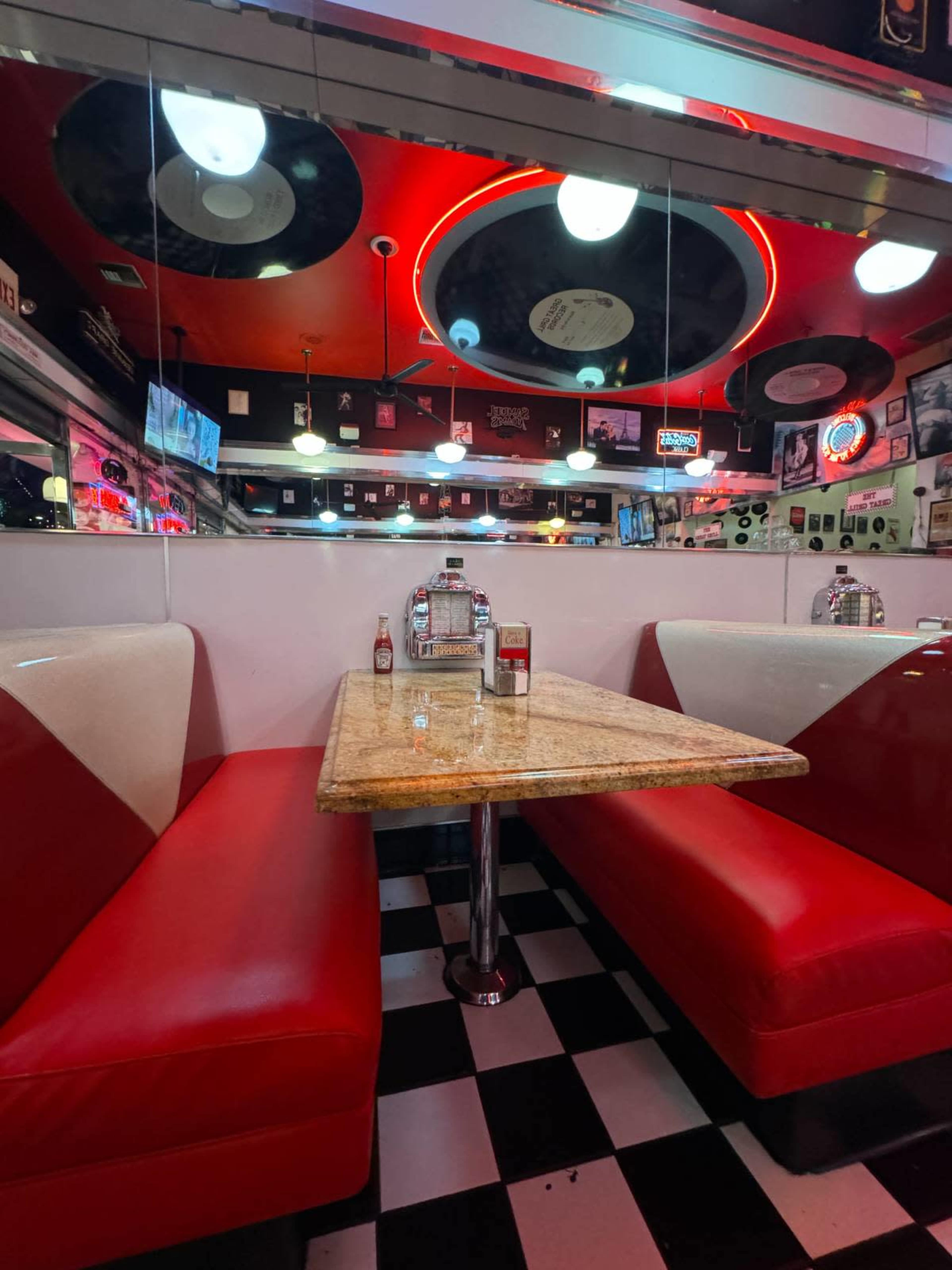 The image shows a retro diner booth with red upholstered seating, a checkered floor, and a mirror-lined wall featuring vinyl records and various memorabilia.