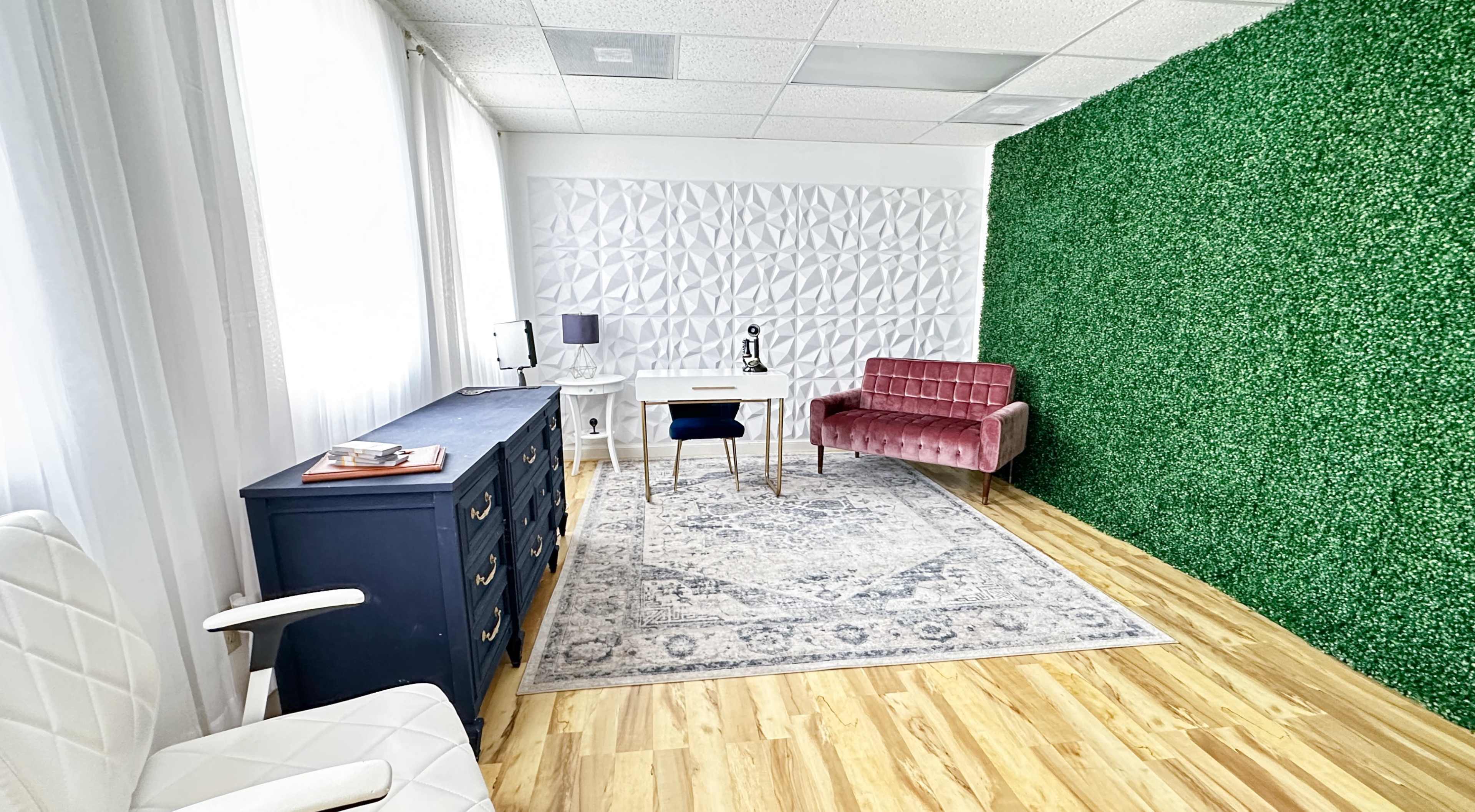 A well-lit office space featuring a green textured wall, a light wooden floor, a white desk with a chair, and a decorative area rug.