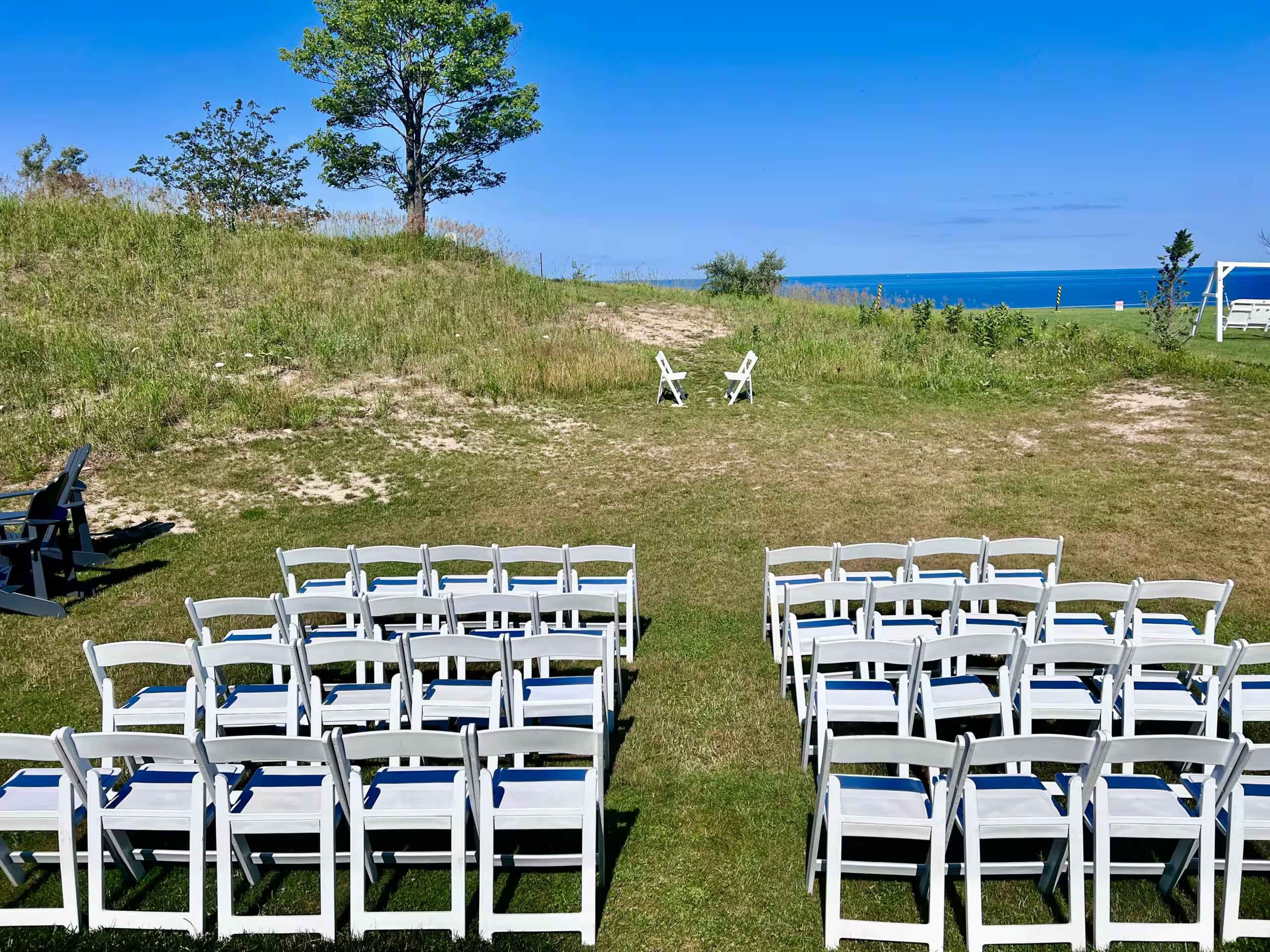 The image shows a grassy area with white folding chairs arranged in rows, facing a distant view of the ocean and two Adirondack chairs on a hill under a clear blue sky.
