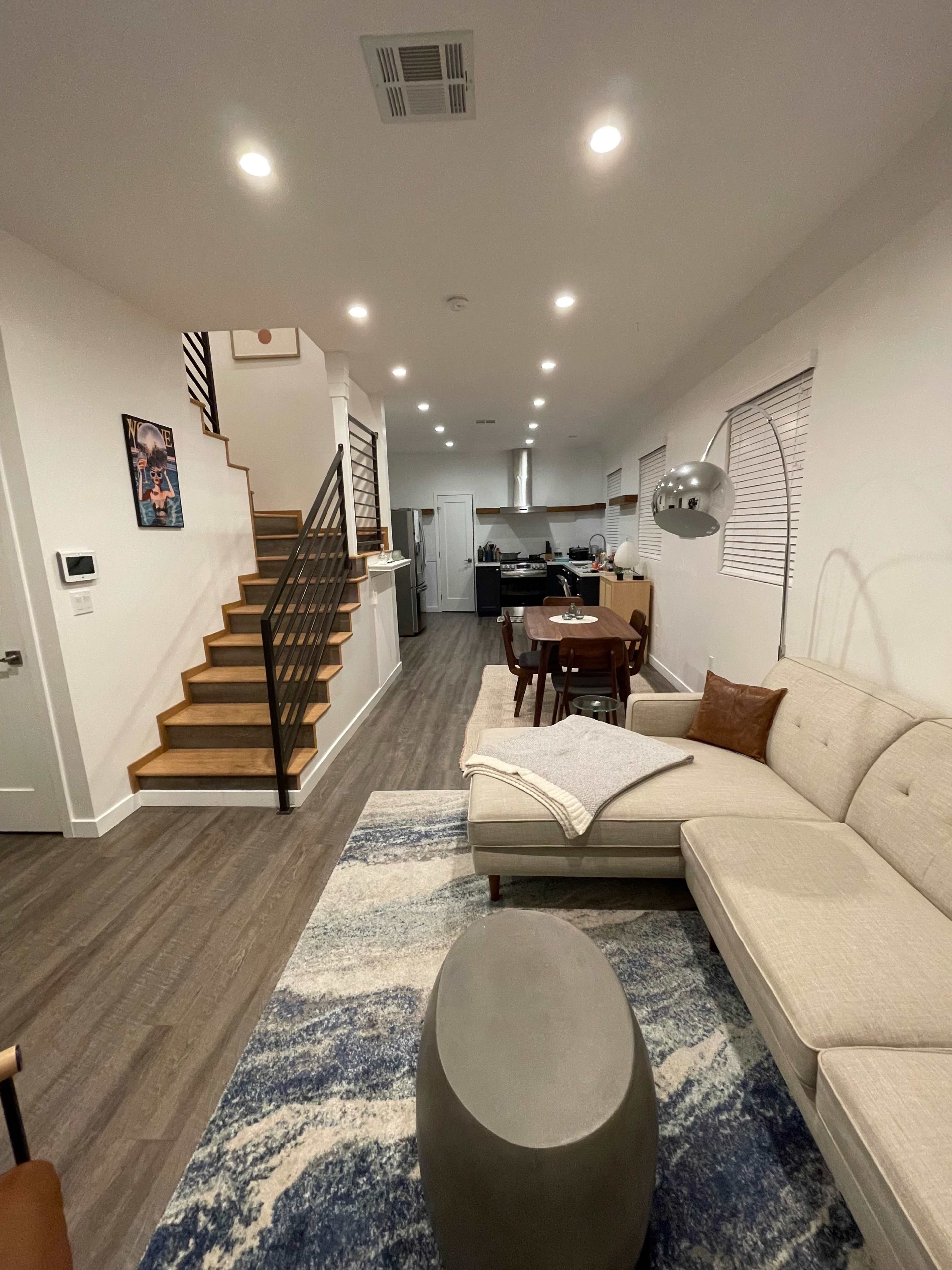 The image shows a modern living area featuring a light-colored sectional sofa, a wooden dining table, and a staircase leading to an upper level, with a kitchen visible in the background.