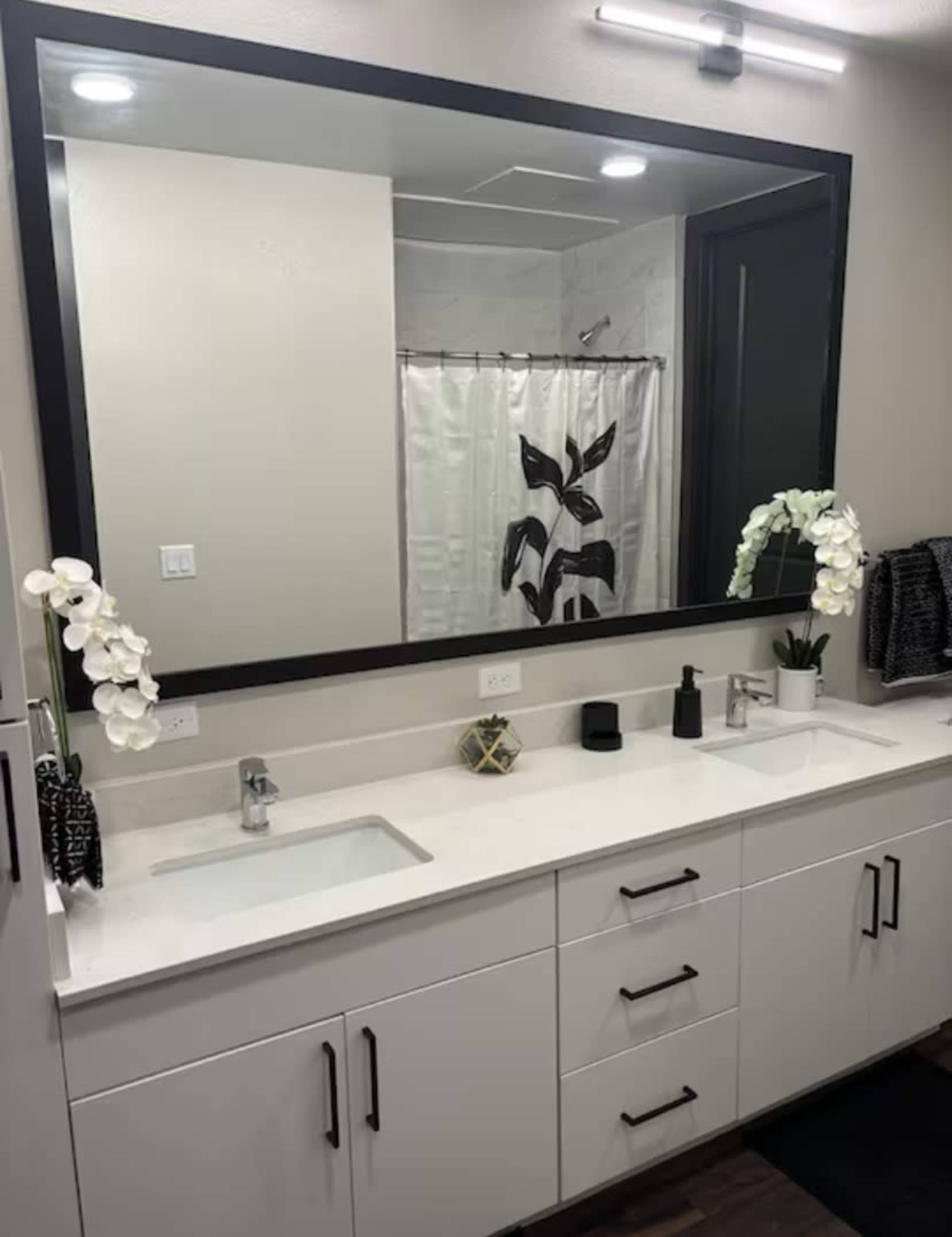 The image shows a modern bathroom with a large mirror, dual sinks, and decorative plants on the countertop.