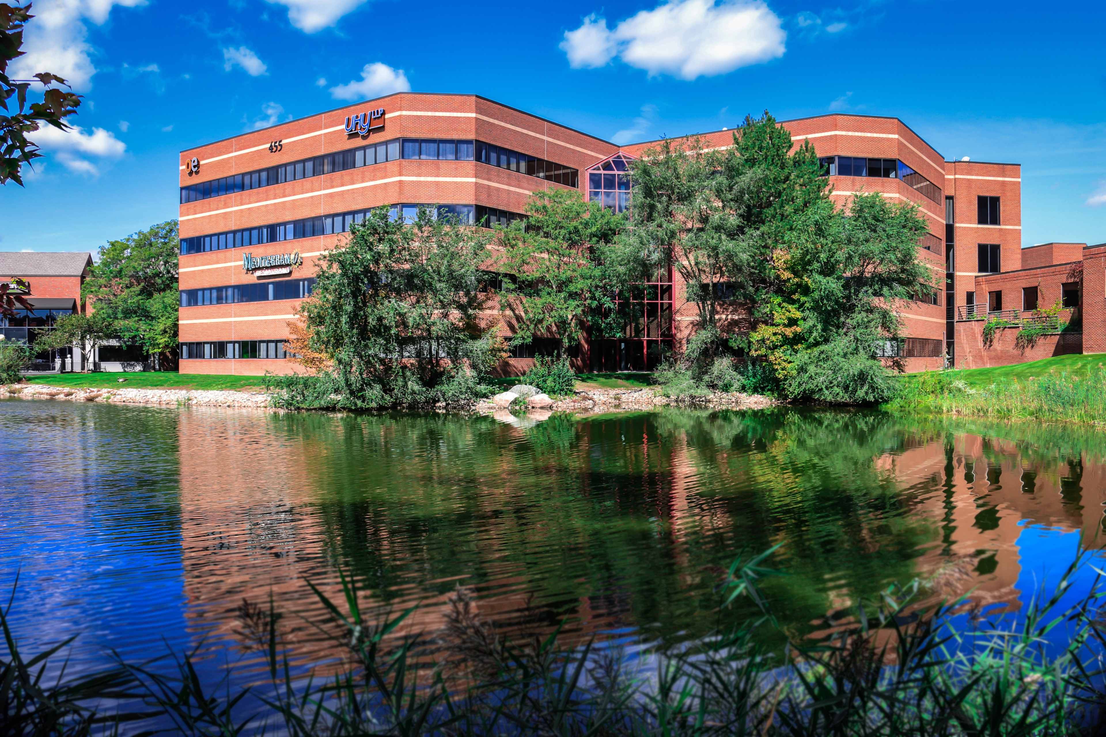 A modern brick building is reflected in a calm pond surrounded by greenery.