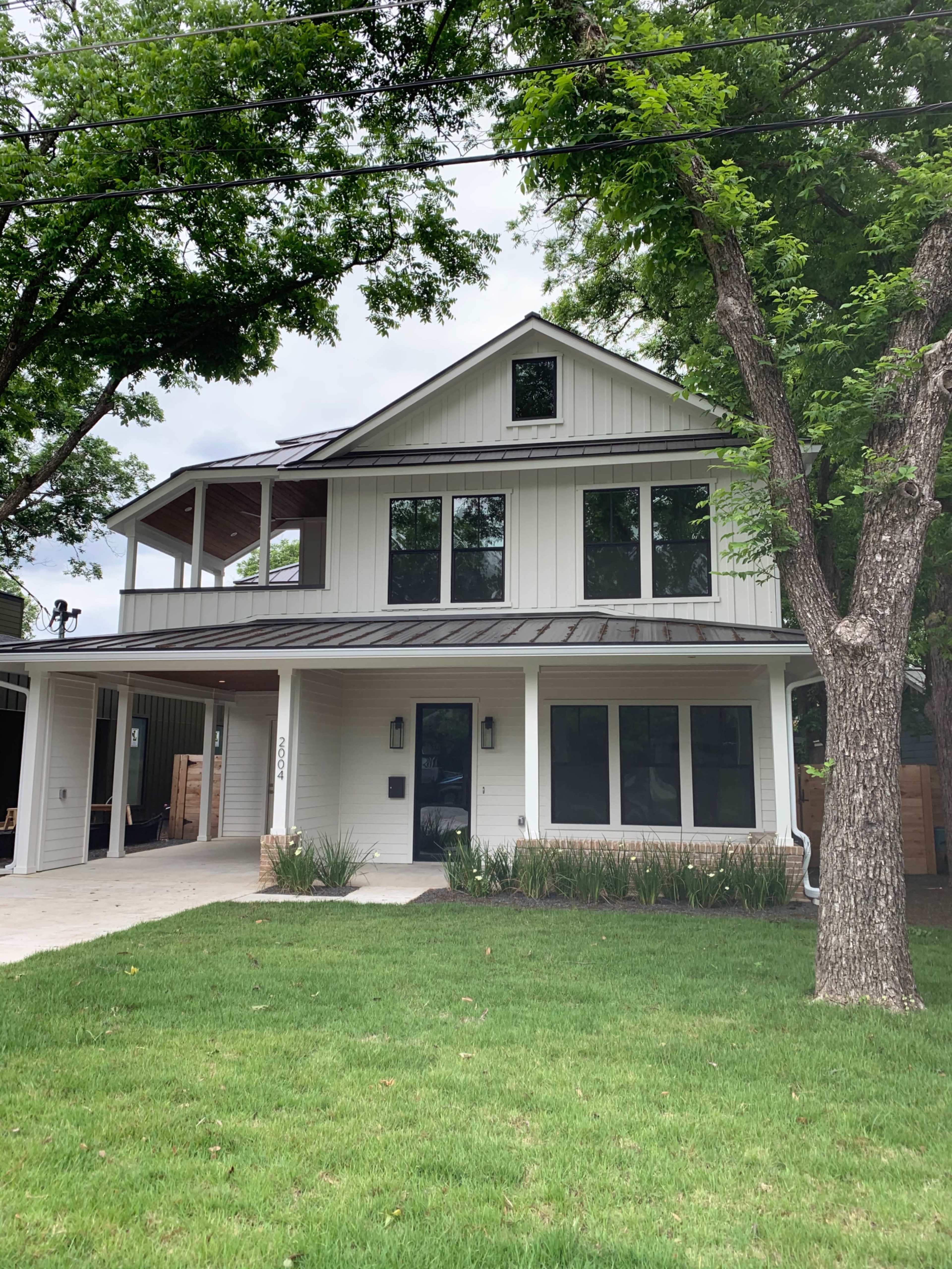 The image shows a modern two-story house with a metal roof, large windows, and a front porch, surrounded by greenery and trees.