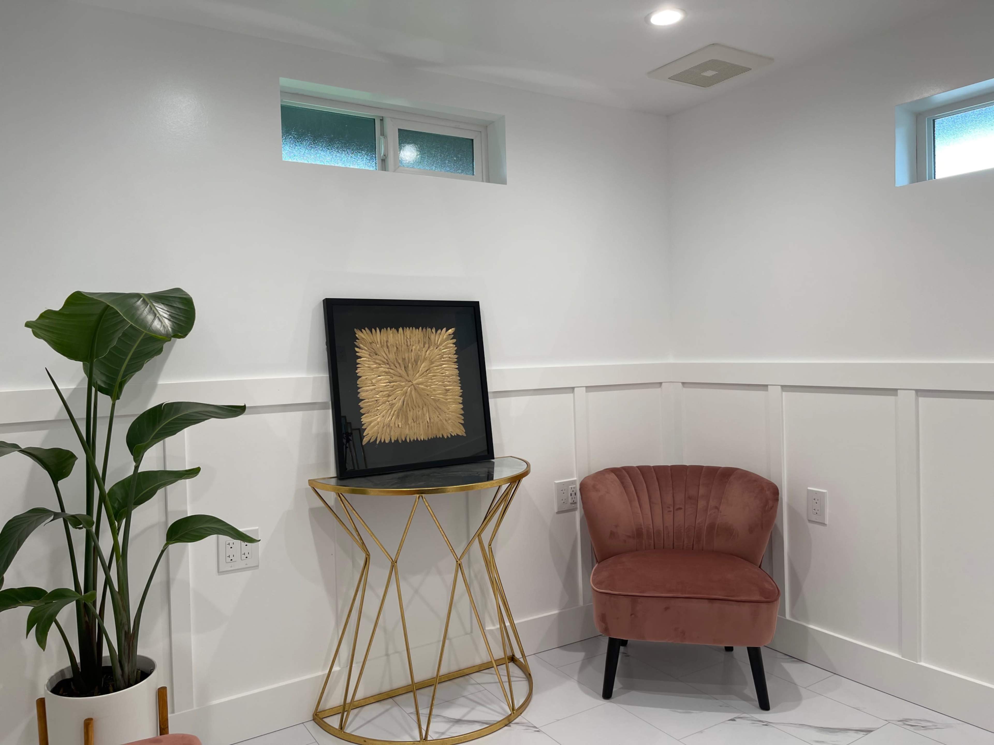 A corner of a modern room featuring a plant, a gold-accented table with a framed artwork, and a pink velvet chair against a white wall.