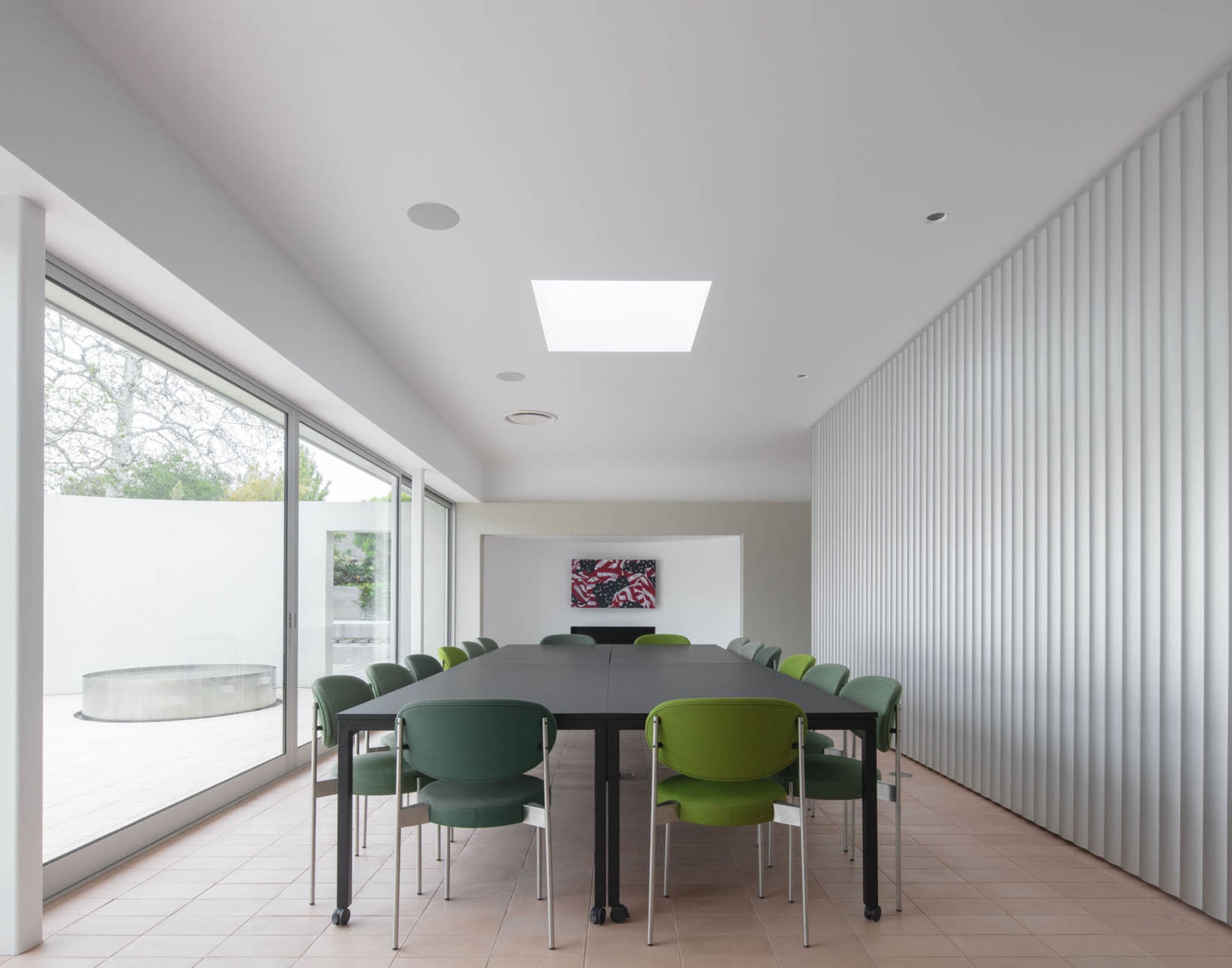 The image shows a modern conference room with a long black table surrounded by green and gray chairs, large glass doors leading outside, and a skylight above.