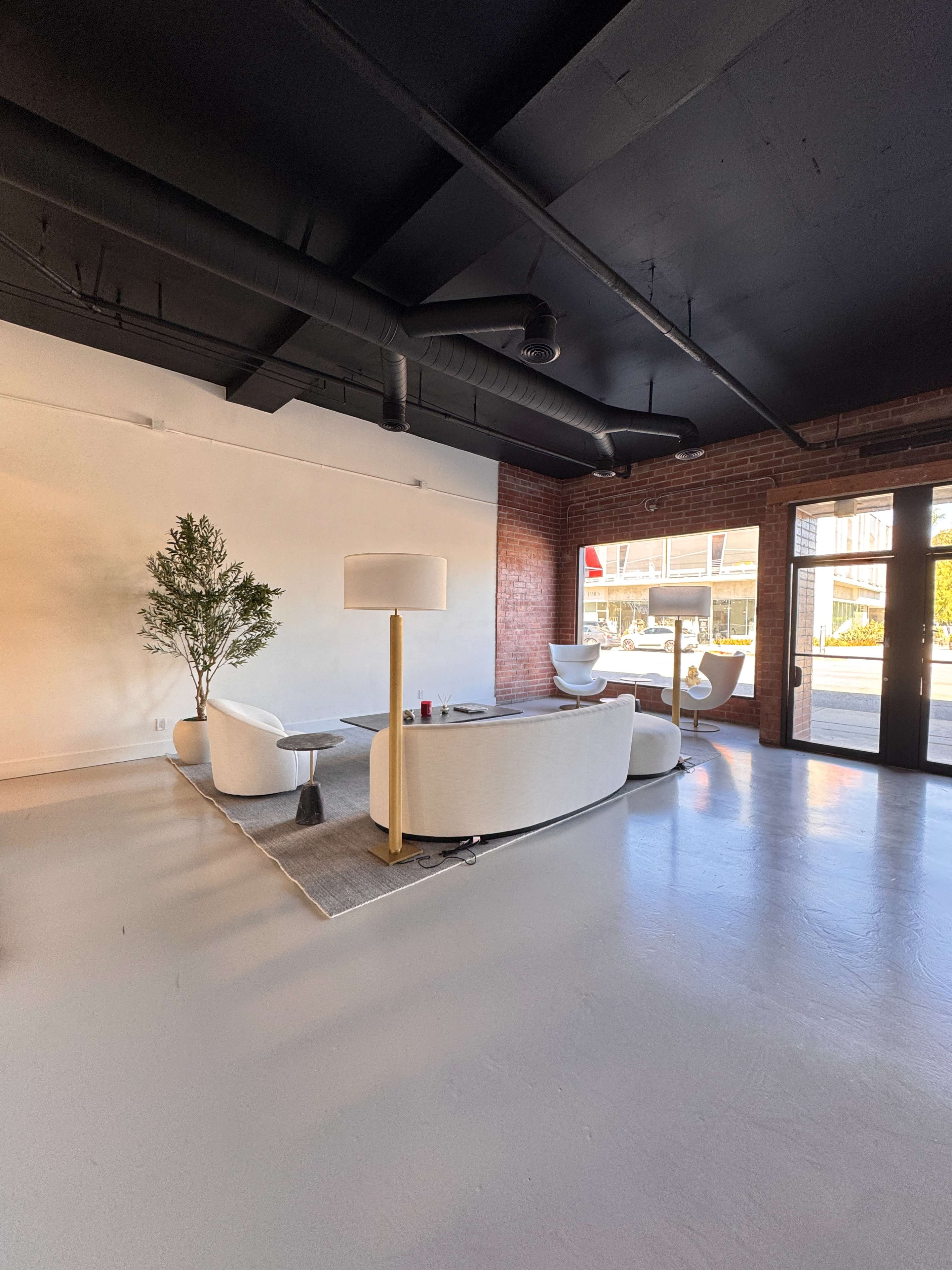 The image shows a modern interior space featuring a white sofa, a lamp, and a small tree, with large windows allowing natural light to enter.