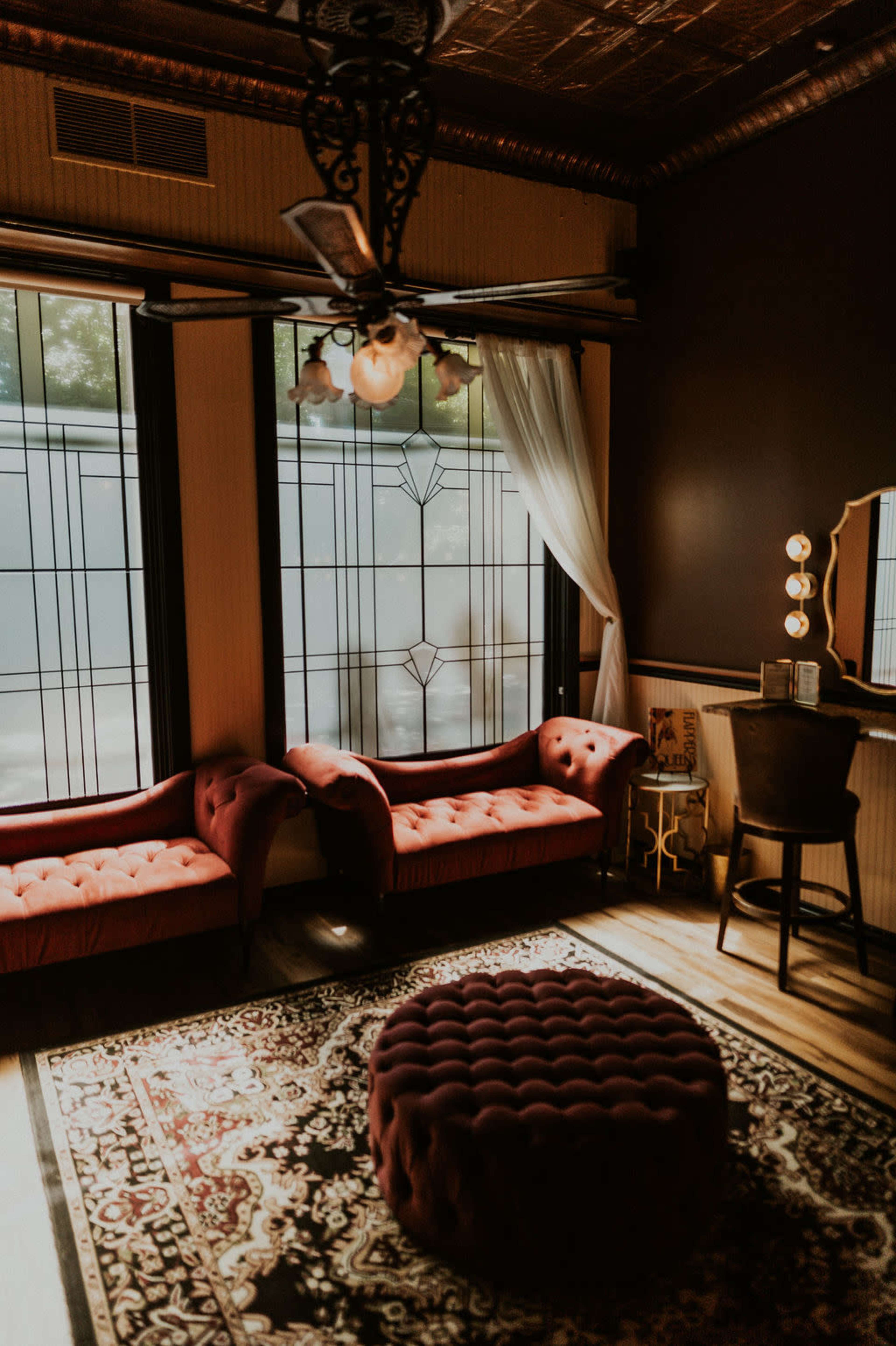 The image shows a cozy room with two tufted red sofas, a round ottoman, and a vanity area illuminated by a mirror with light bulbs, surrounded by large windows.