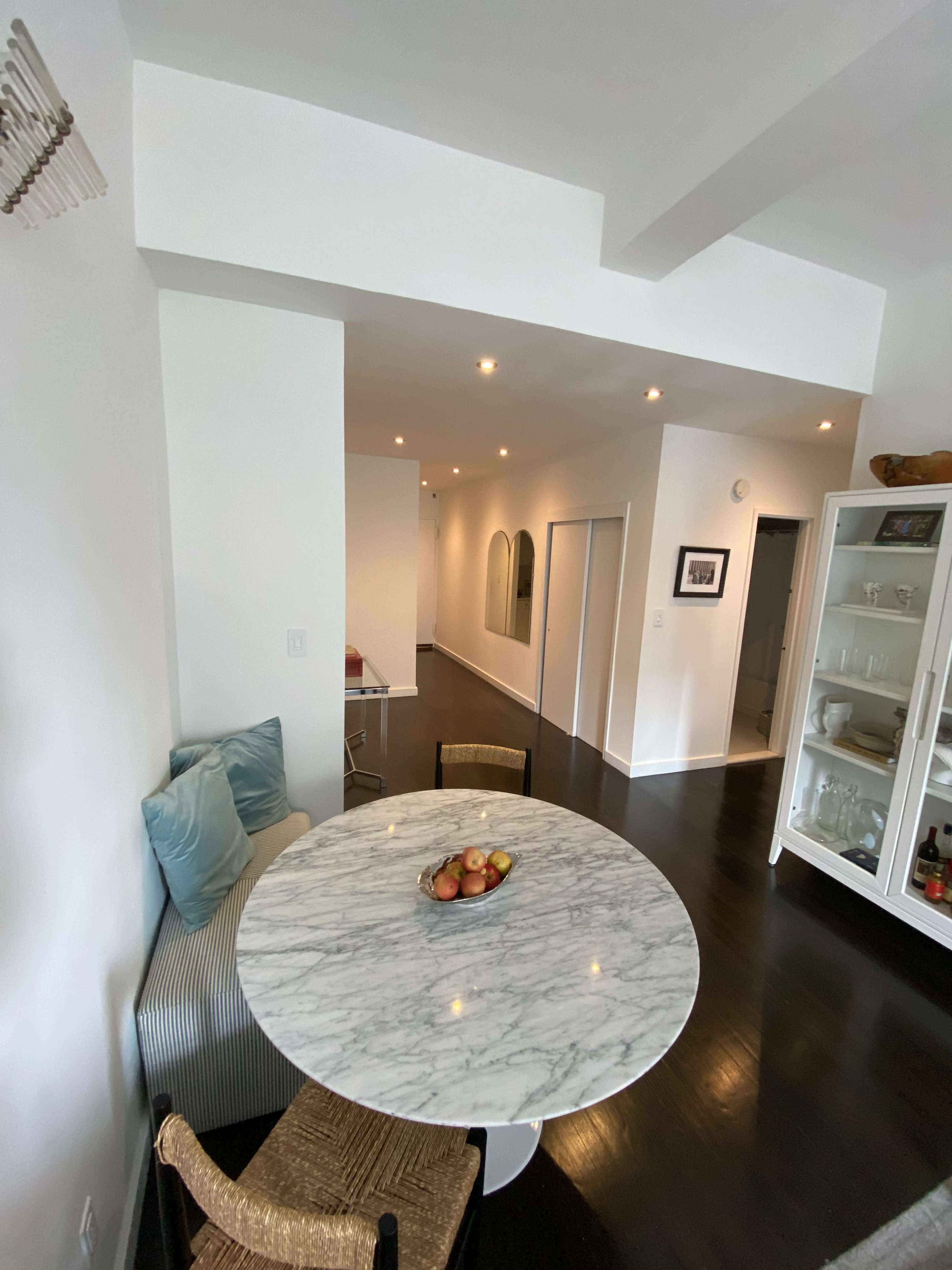 A round marble table with a fruit bowl is set in a bright, minimalistic dining area that leads to a long hallway.