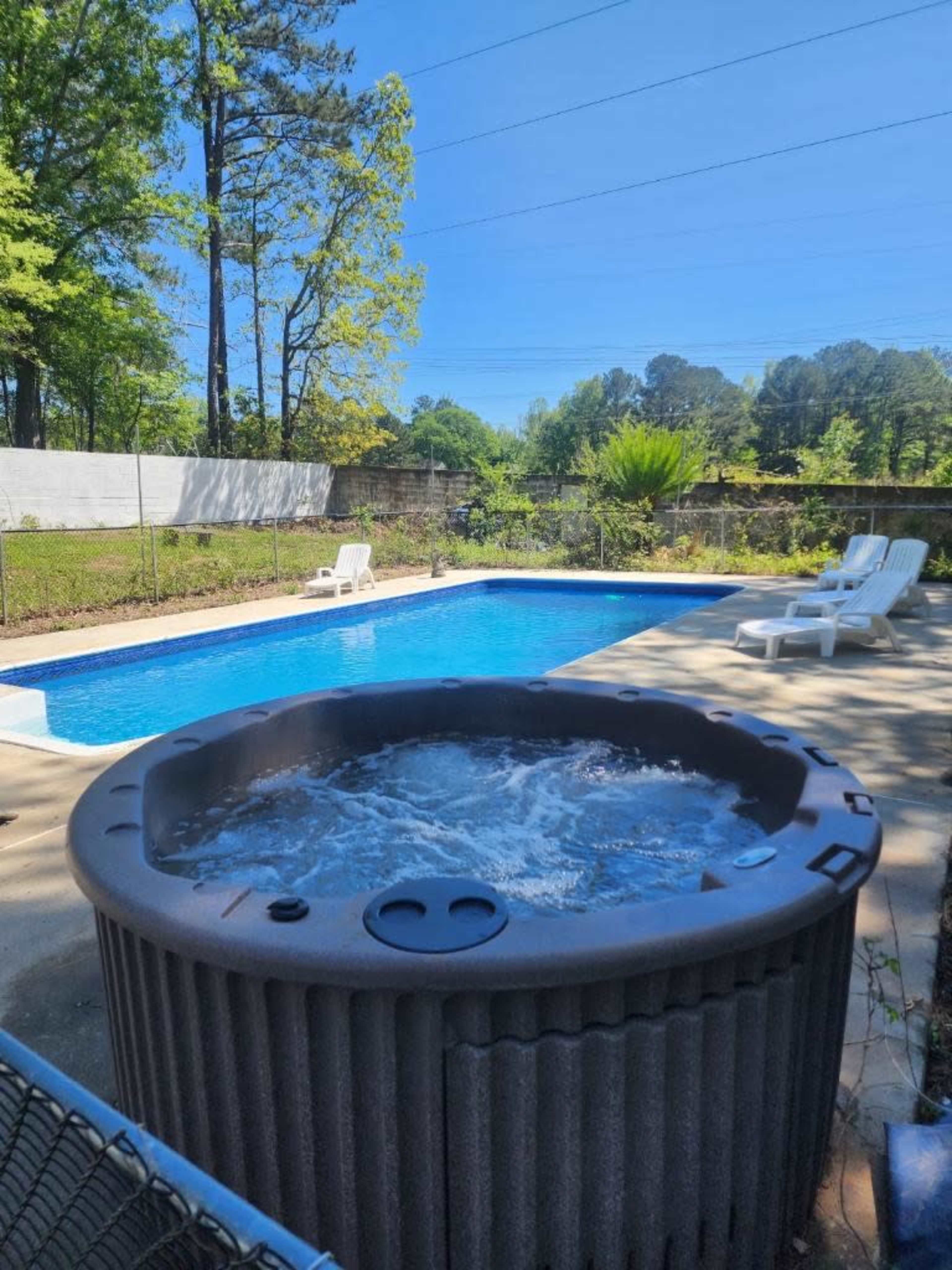 A hot tub is positioned next to a clear blue swimming pool surrounded by green trees and lawn chairs.