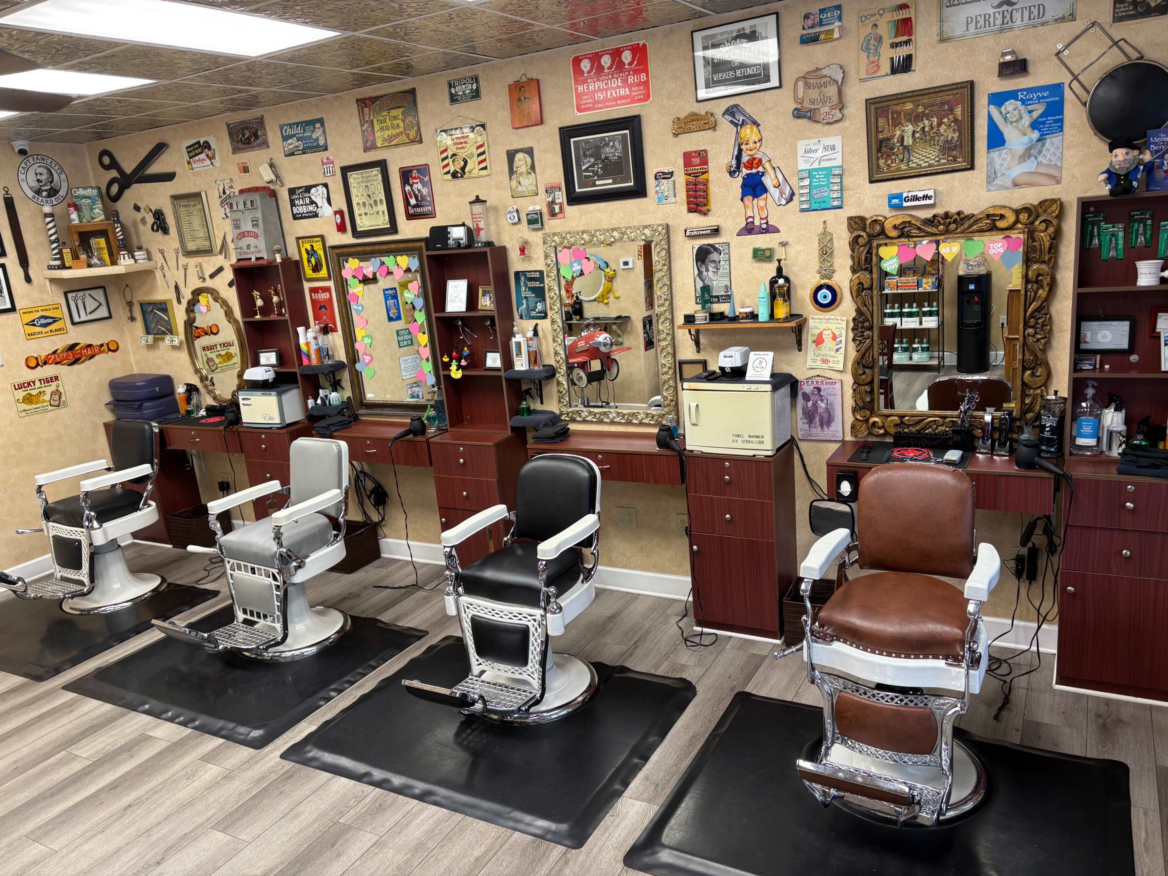 The image shows a barbershop interior with four vintage-style barber chairs arranged in front of mirrors, surrounded by a wall adorned with various signs and decorations.