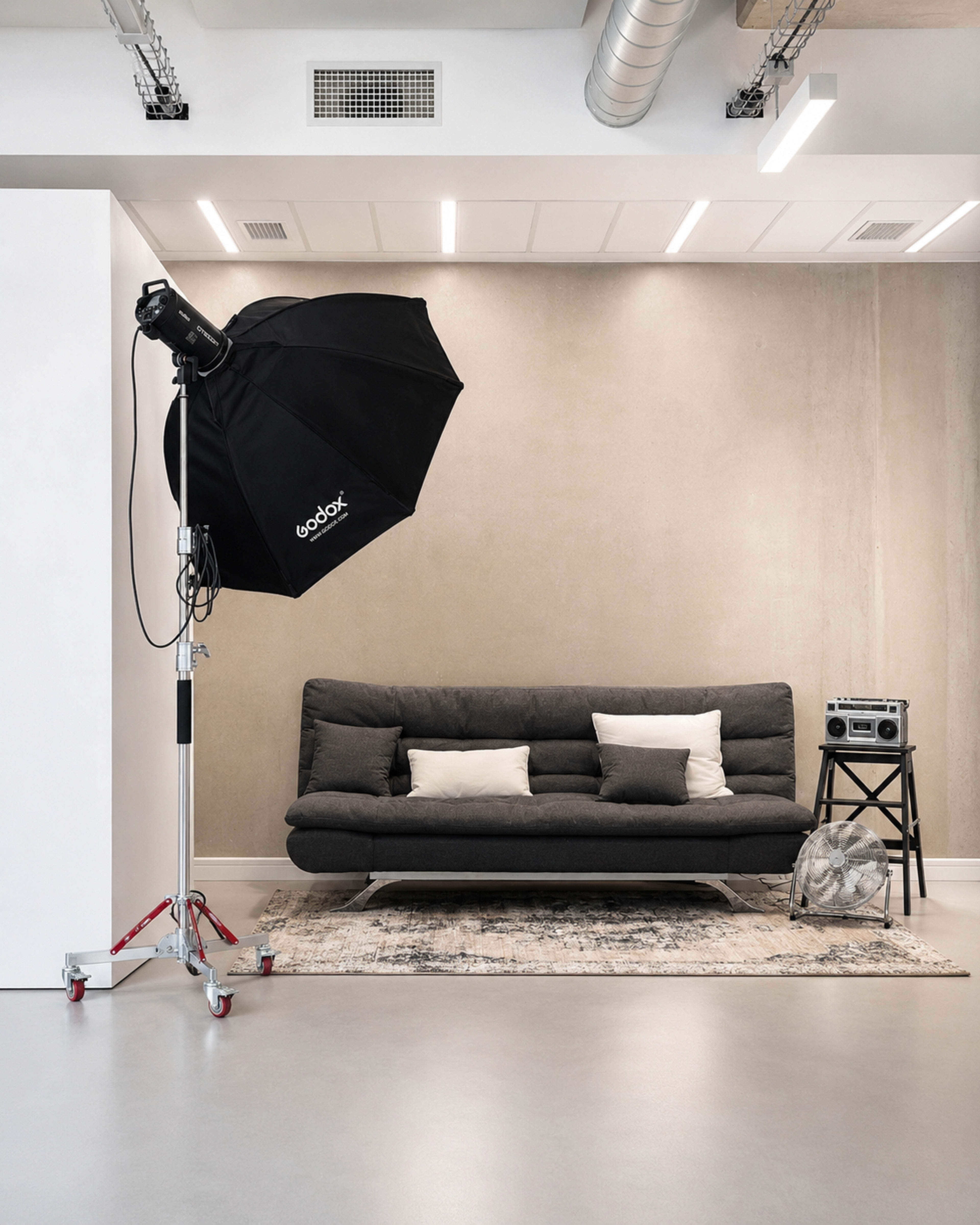 The image shows a minimalist interior with a gray sofa, a side table holding a vintage speaker, a spotlight on a stand, and a textured wall.