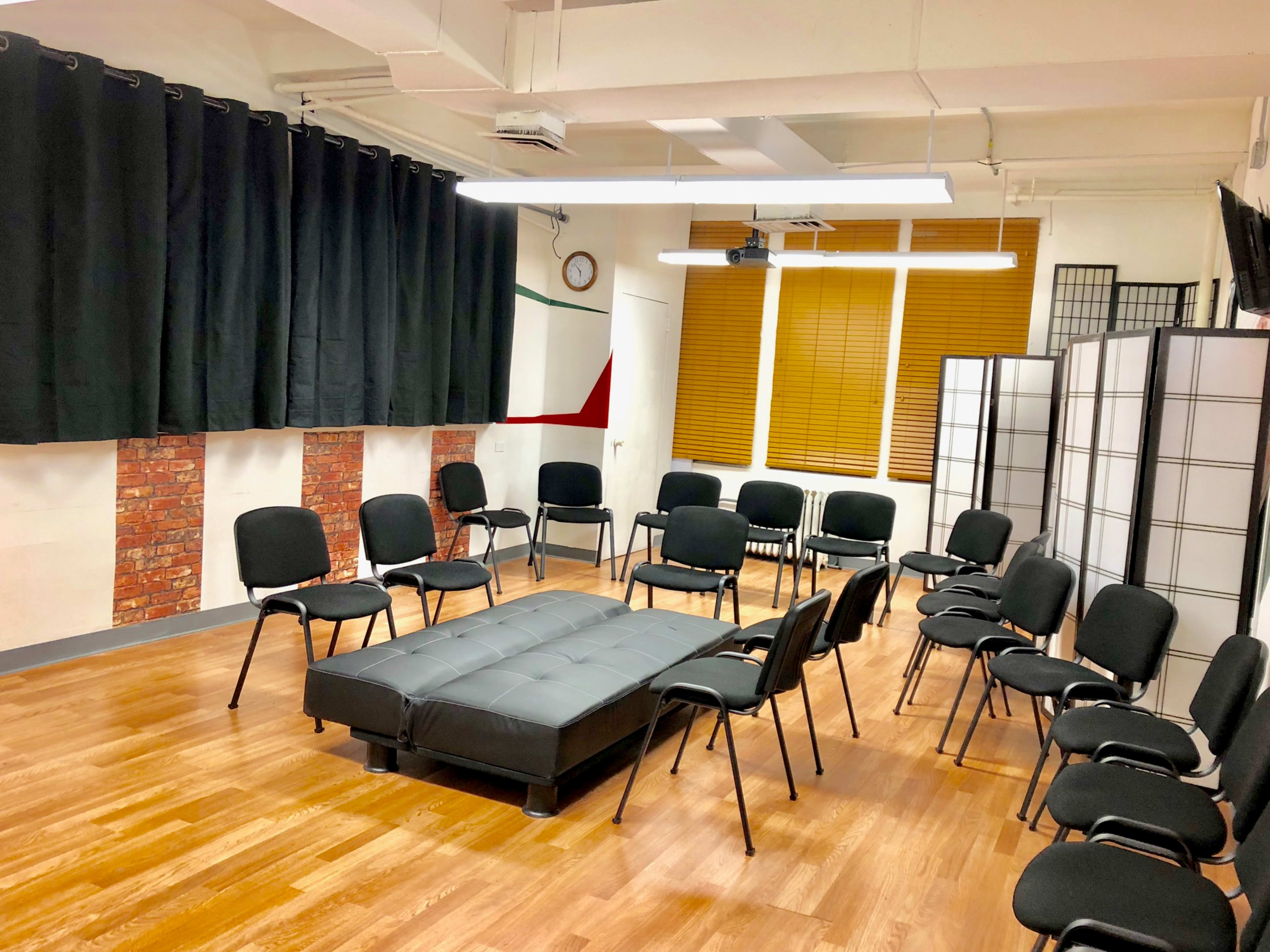 A room arranged with a circular seating layout of black chairs around a dark sofa on a wooden floor, with black curtains and a folding screen in the background.