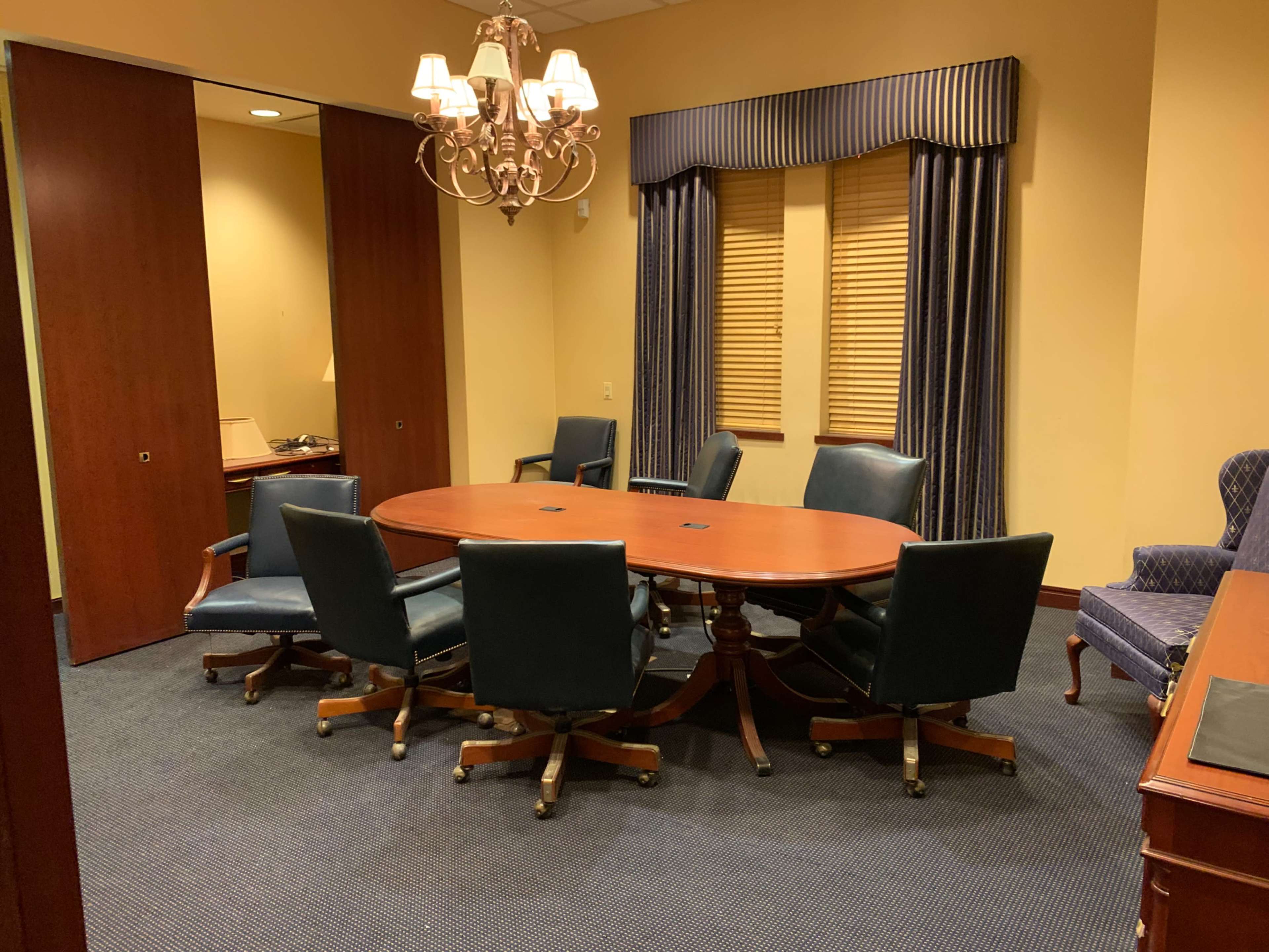 The image shows a conference room with a large round wooden table surrounded by six blue leather chairs, featuring a chandelier and window treatments.