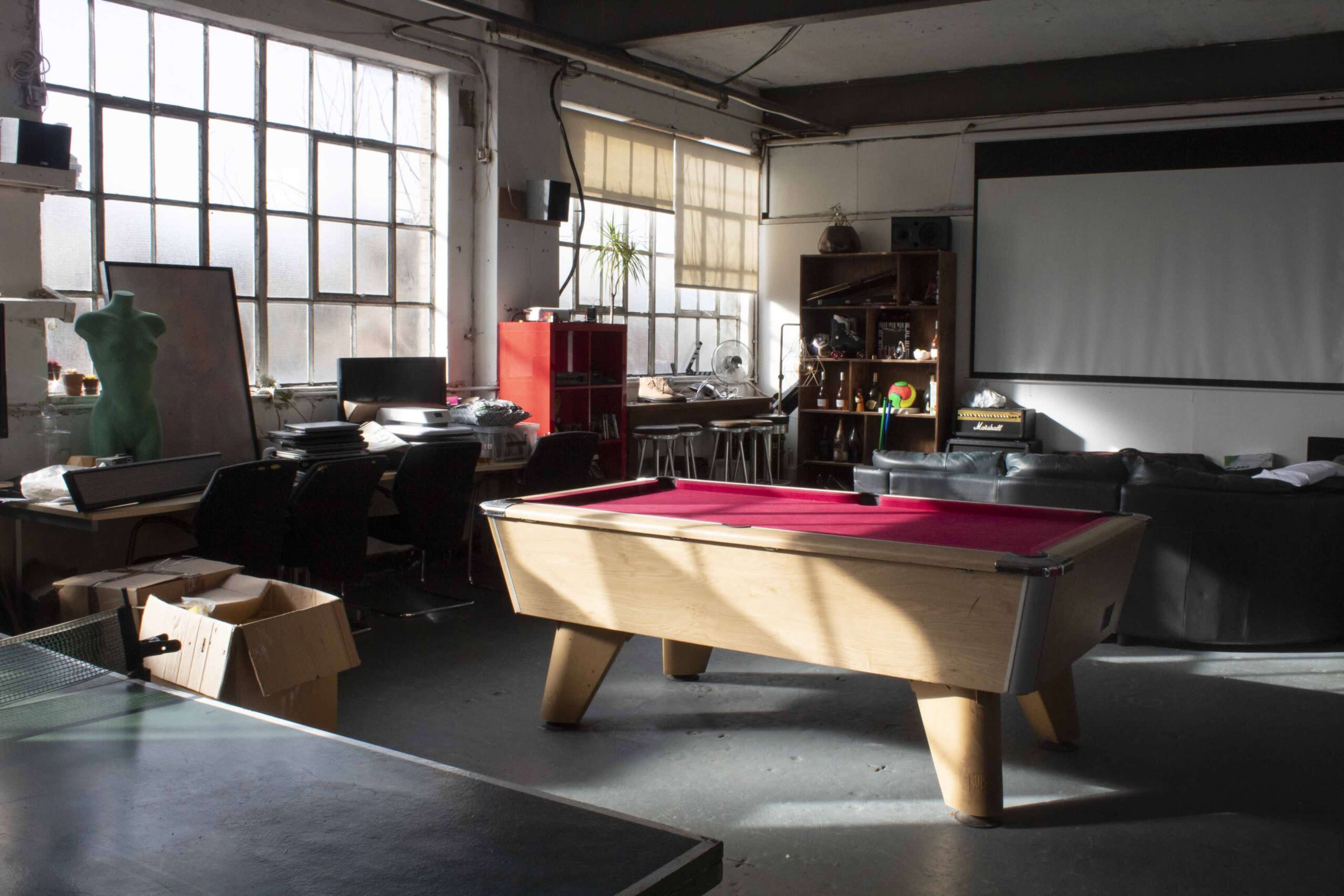 An industrial-style room with a pool table in the center, surrounded by furniture, shelves, and large windows that allow natural light to enter.