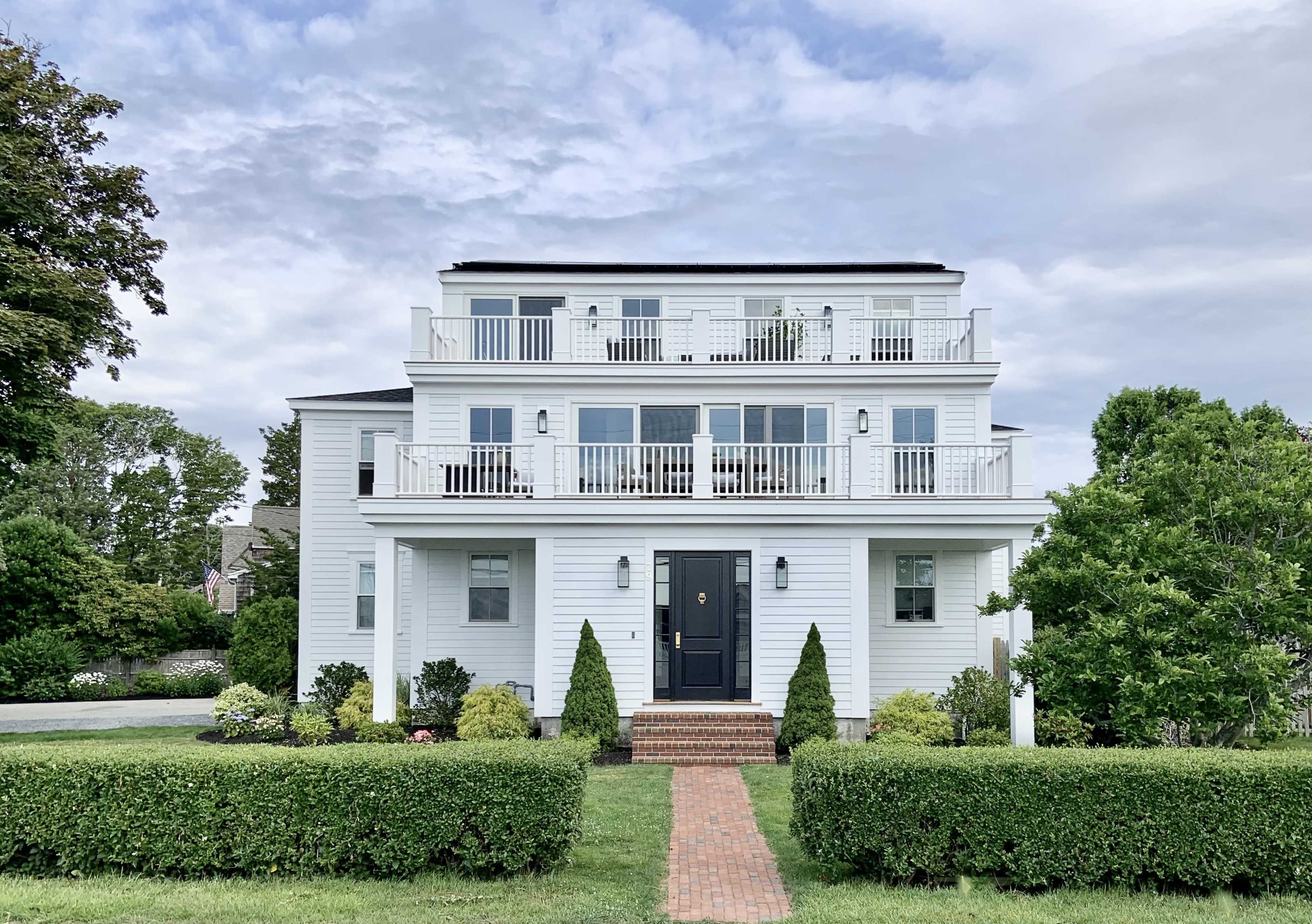 A three-story white house with multiple balconies and a brick walkway leads to the front door, surrounded by neatly trimmed hedges and green landscaping.