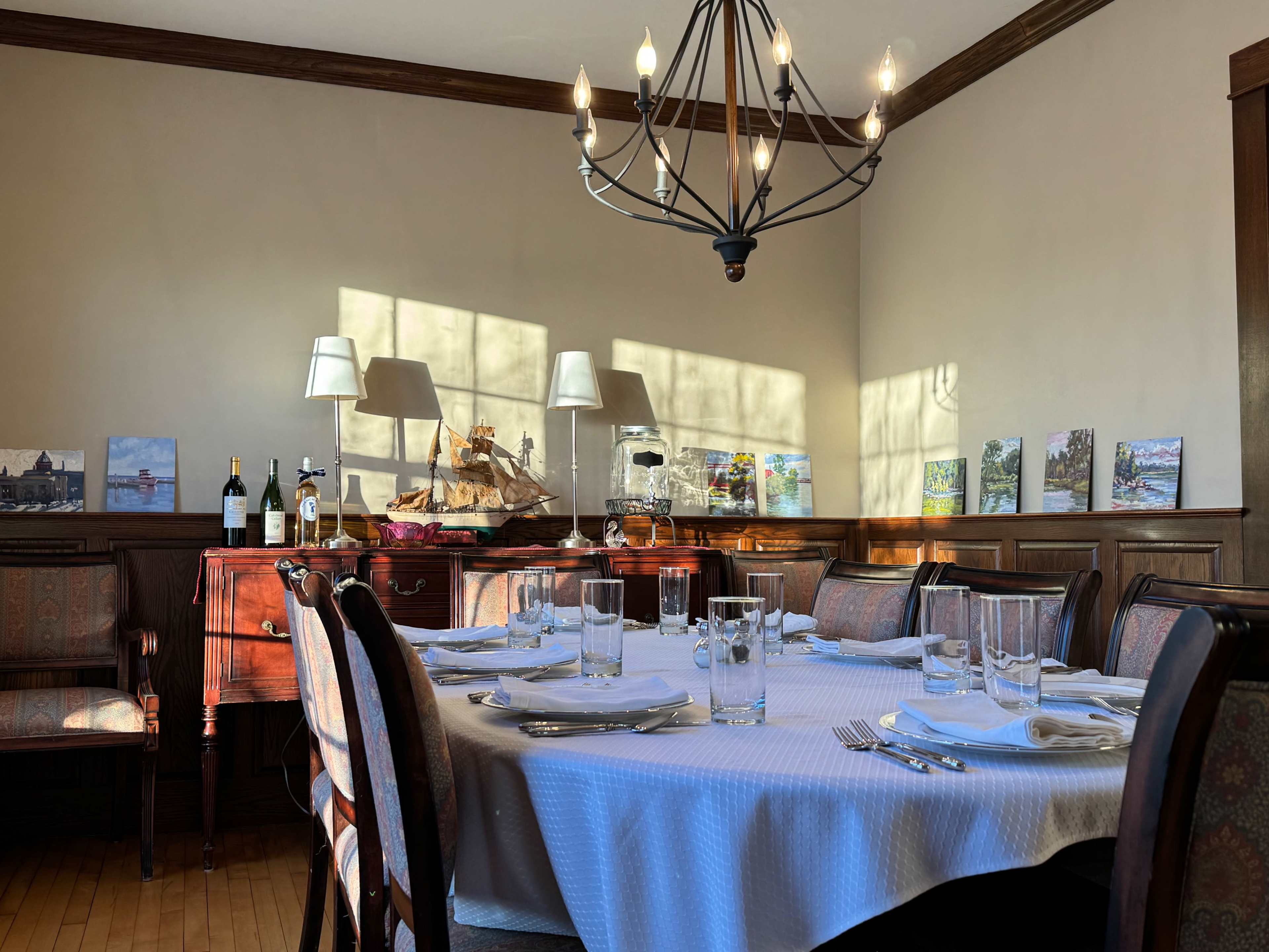 A dining room with a set table and a chandelier, featuring a sideboard with wine bottles and framed artworks on the wall.