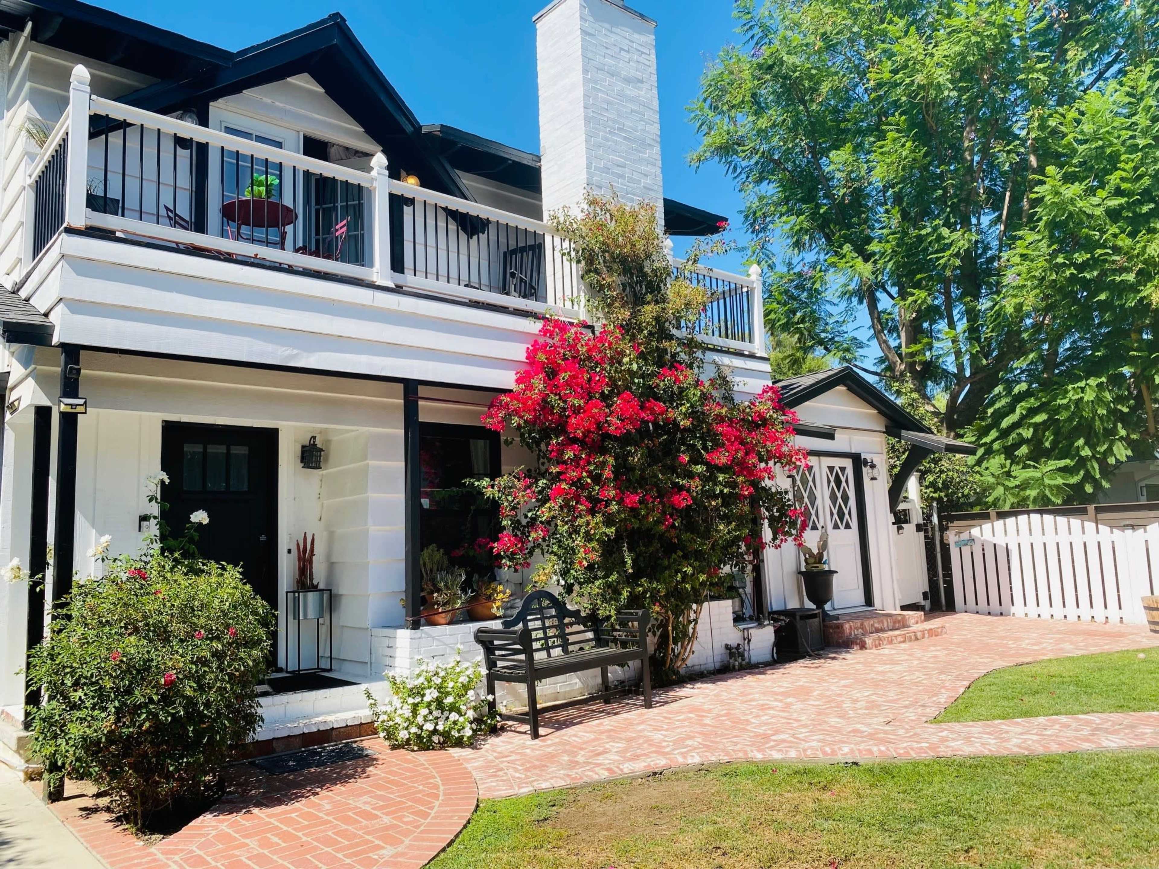 A two-story house with a white exterior, a brick pathway, and a front garden featuring blooming flowers and a bench.