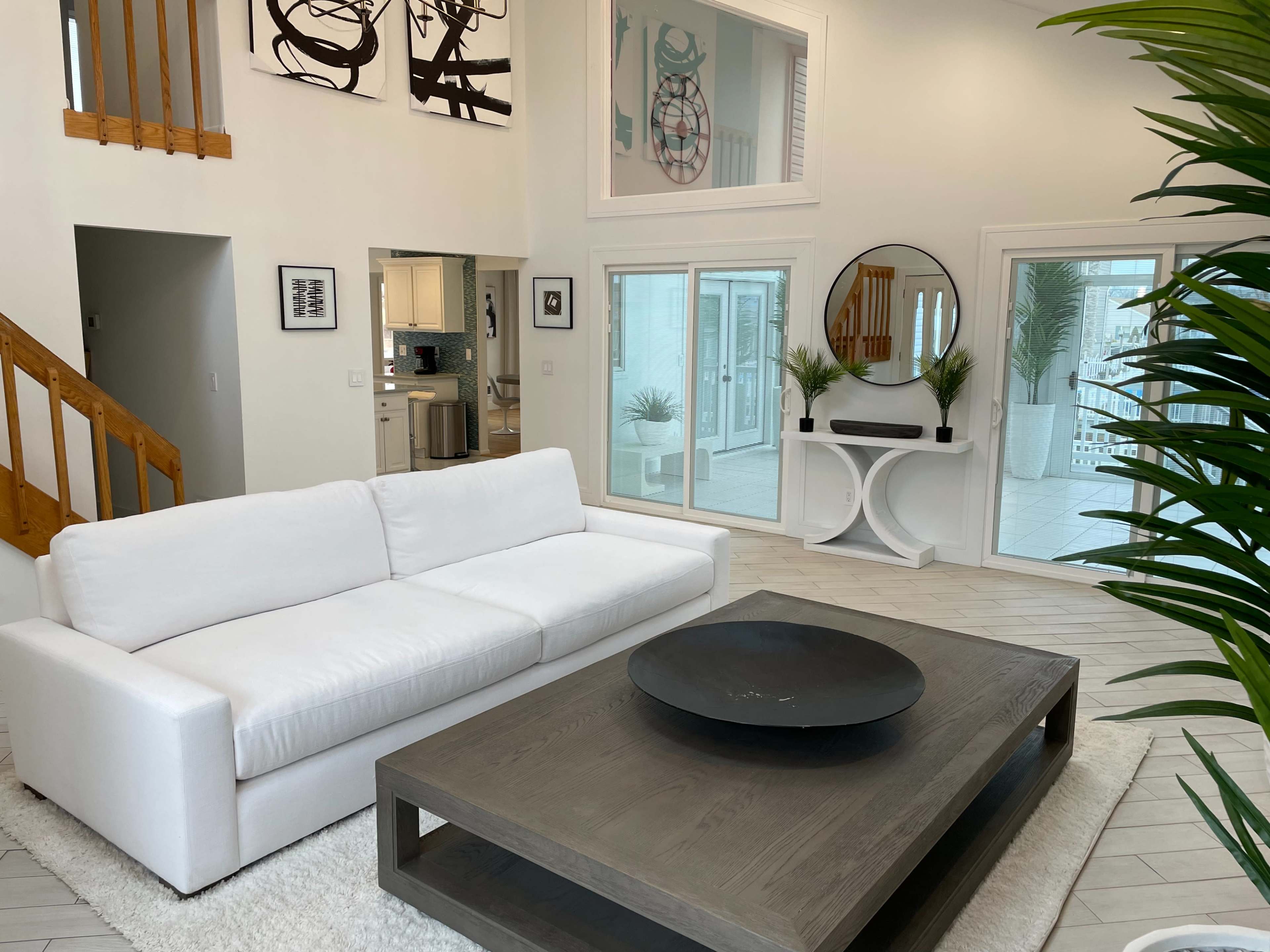 The image shows a modern living room featuring a white sofa, a large rectangular coffee table, and a bright, open layout with large windows and a staircase in the background.
