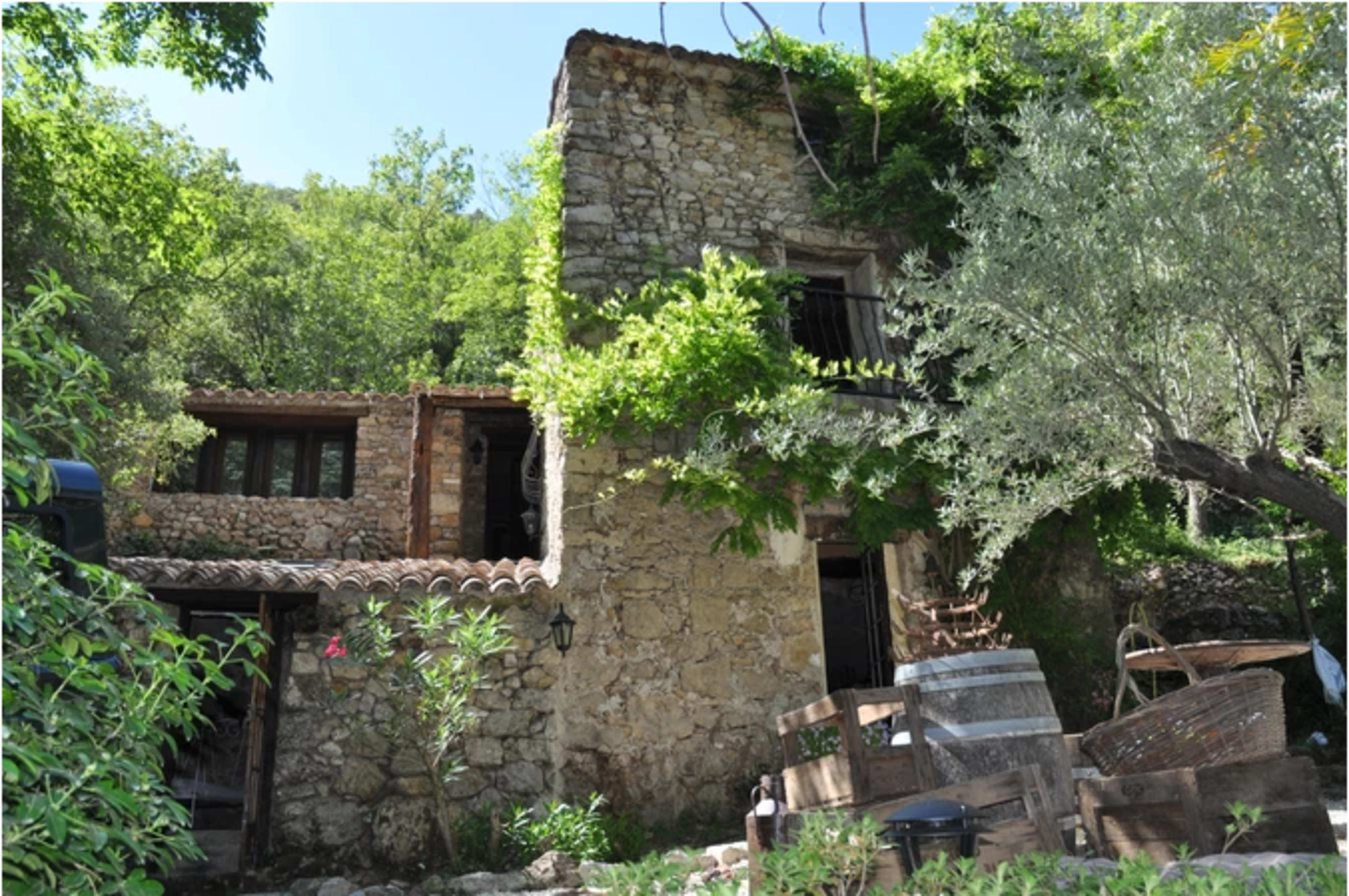 A rustic stone house is surrounded by greenery, with vines climbing the walls and wooden barrels positioned in the foreground.