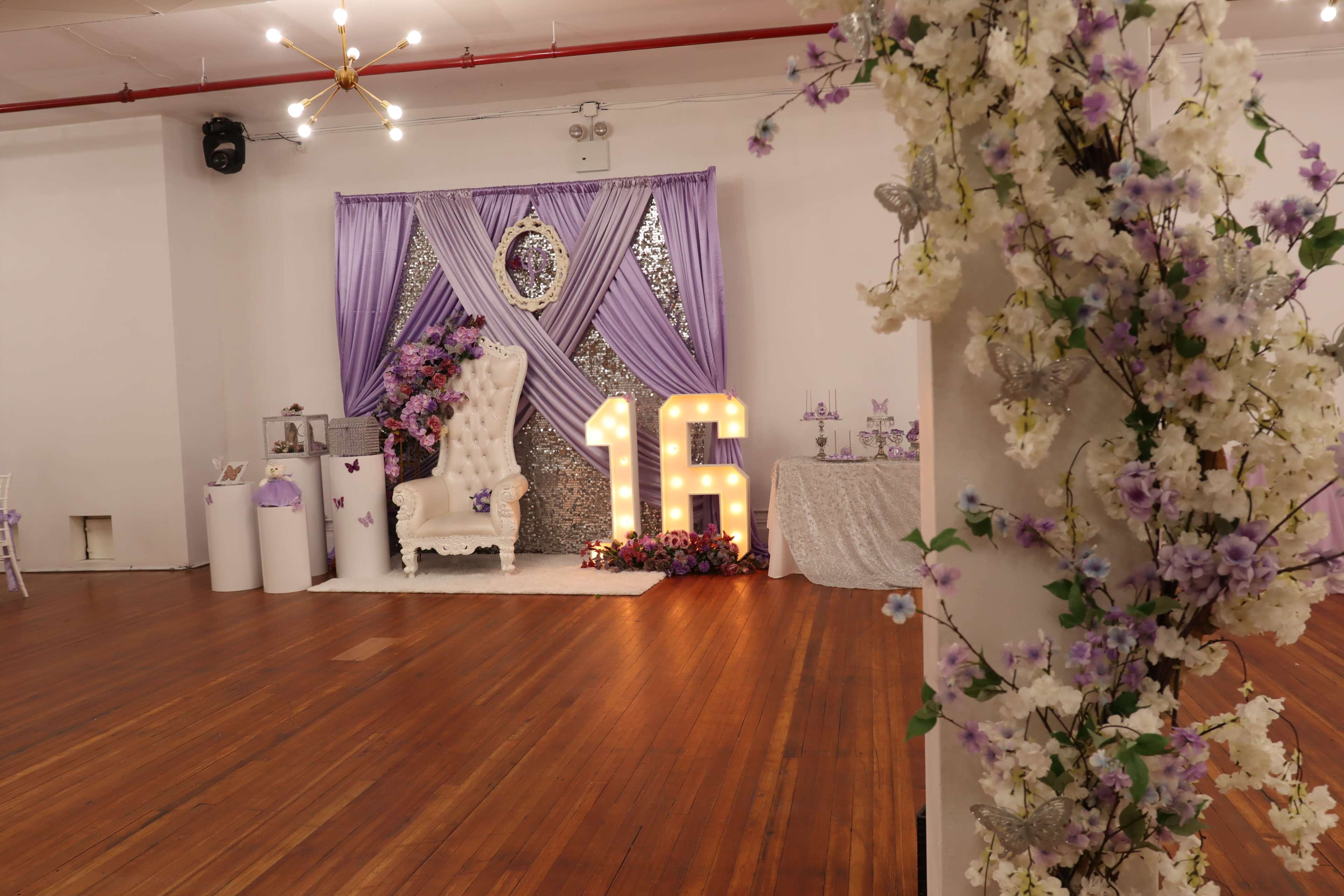 A party setup featuring a large "16" display, a decorated backdrop with purple drapery, and floral arrangements in a spacious indoor venue.