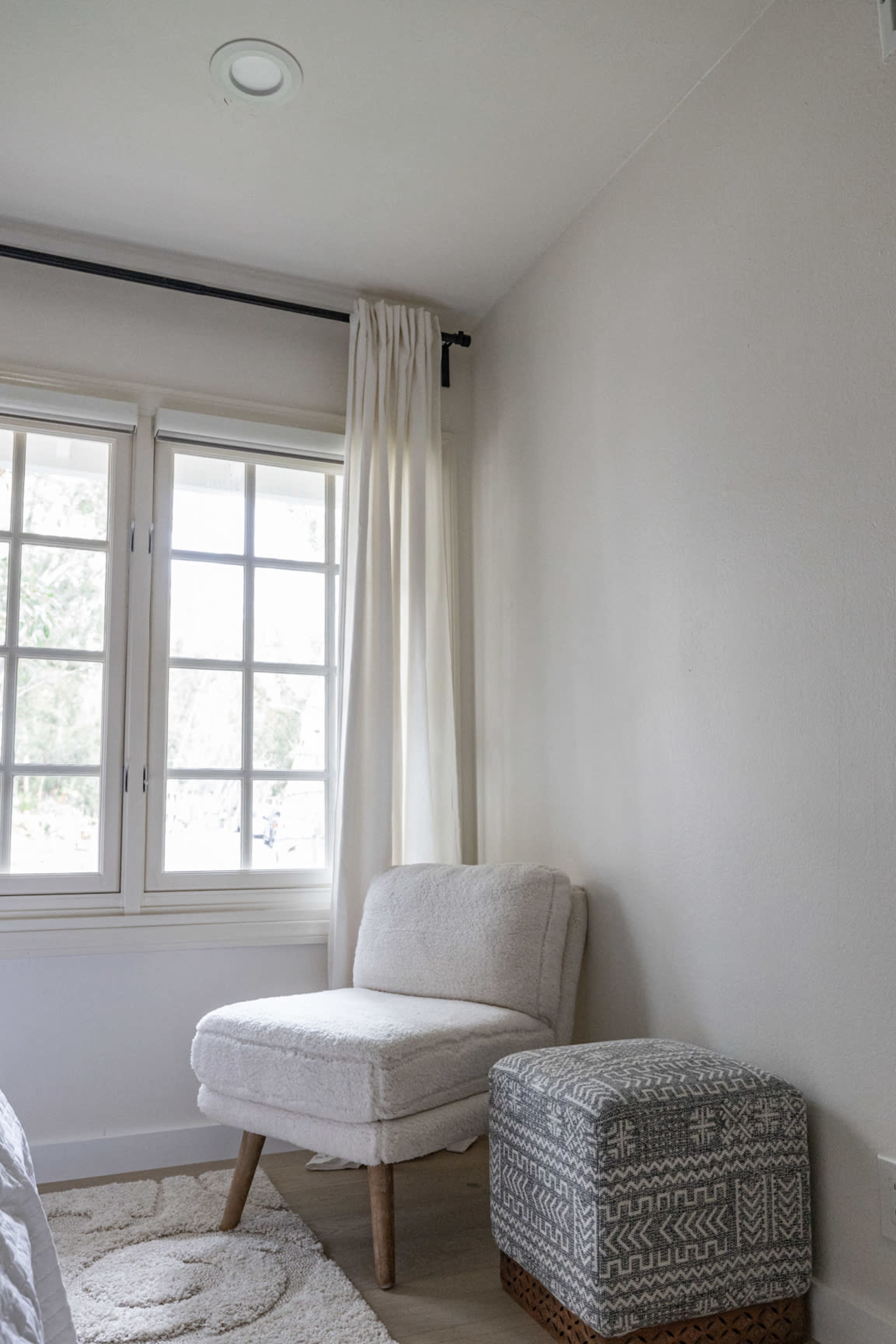 A light-colored chair and a patterned ottoman are positioned next to a large window with sheer curtains in a softly lit room.