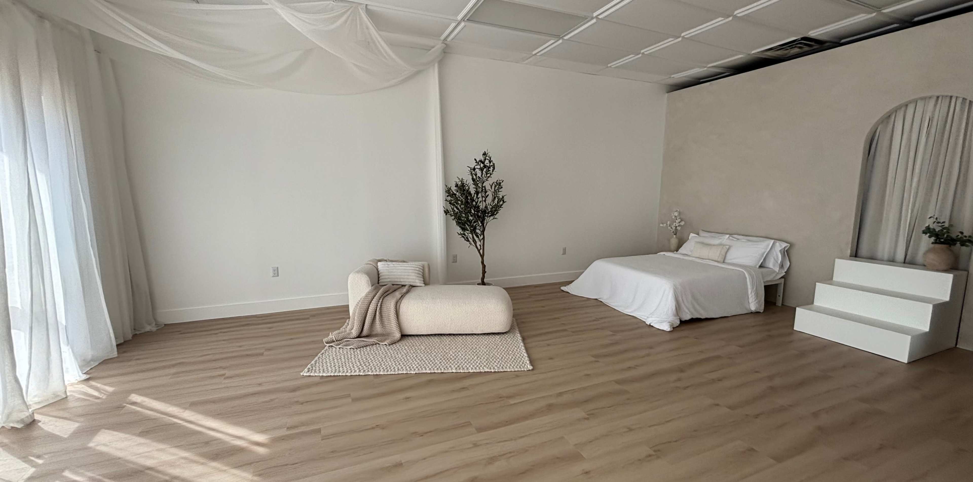 The image depicts a minimalist bedroom with a bed, a round cushion, a woven rug, and a large window allowing natural light to enter.