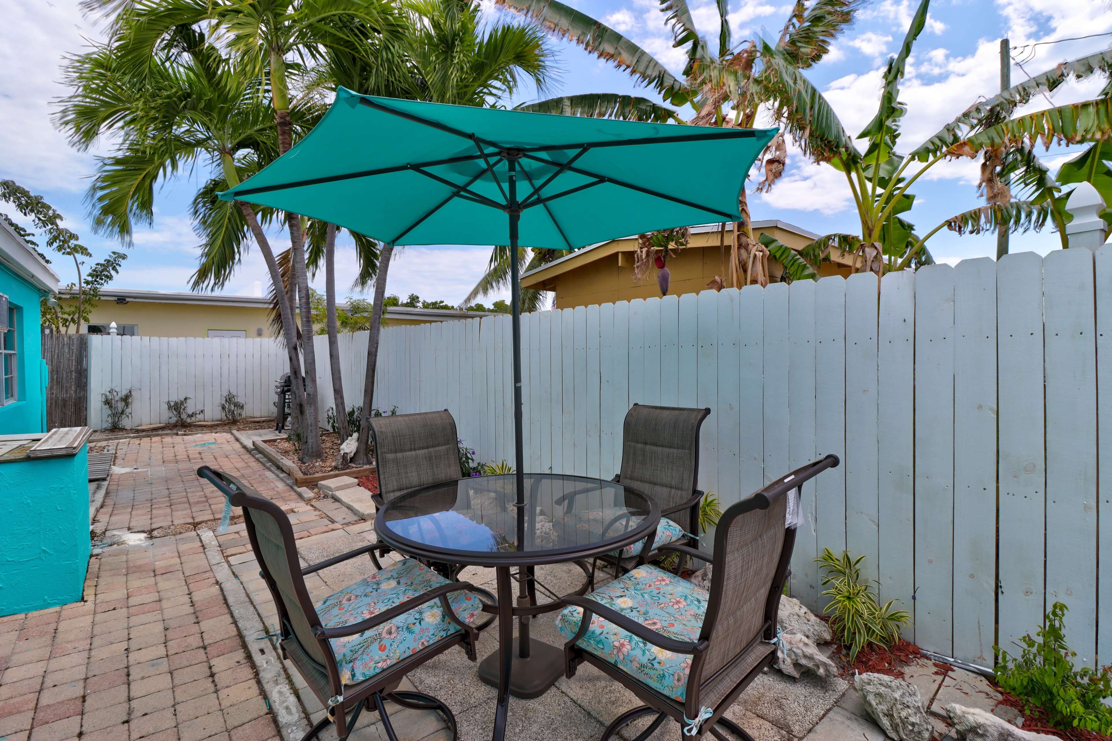 A patio area features a round table with four chairs under a teal umbrella, surrounded by palm trees and a white picket fence.