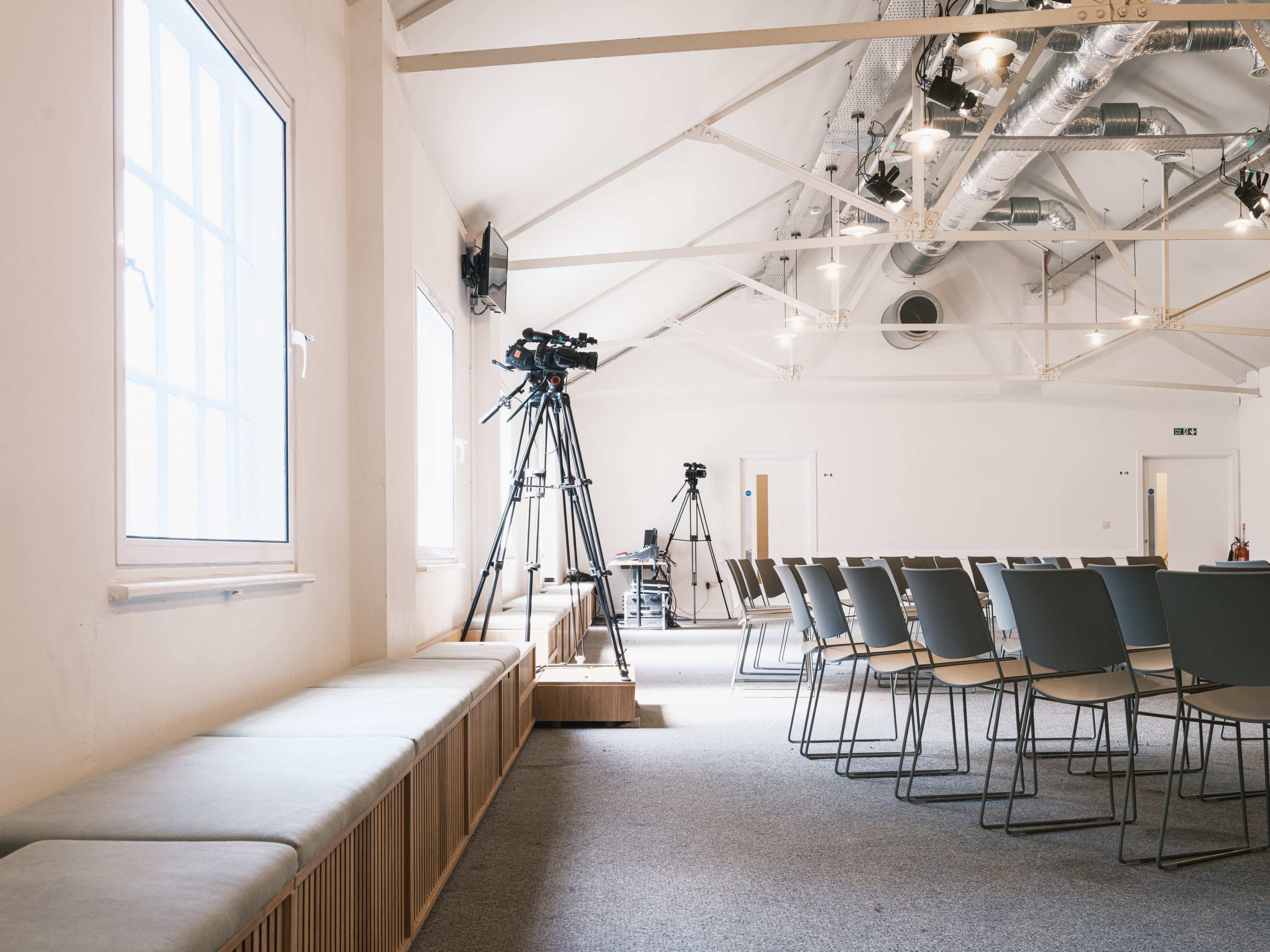 A spacious room with rows of chairs arranged in front of a video camera setup and large windows providing natural light.