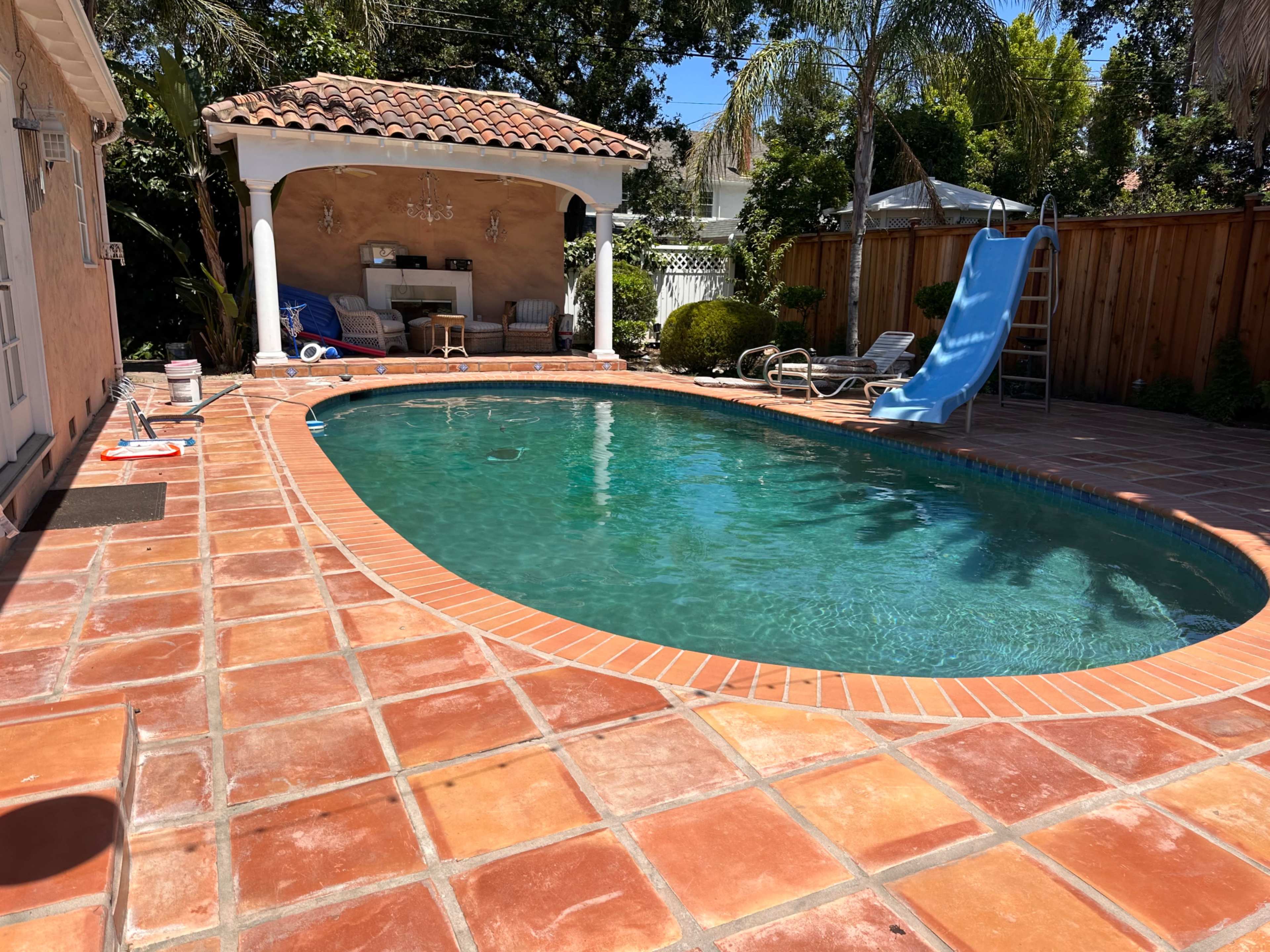 A swimming pool with a tiled deck is surrounded by a wall and features a shaded seating area and a ladder leading into the water.