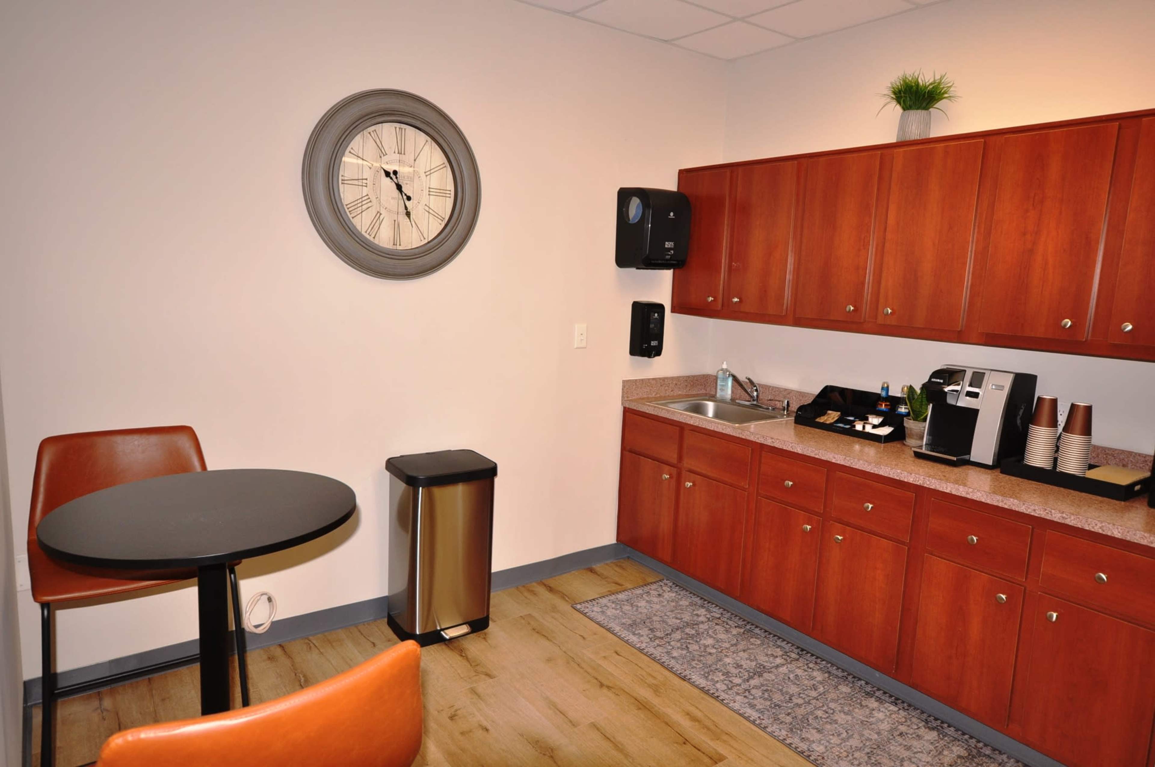 The image shows a break room featuring a small round table with two orange chairs, a clock on the wall, a waste bin, and a kitchenette with wooden cabinets and a coffee machine.