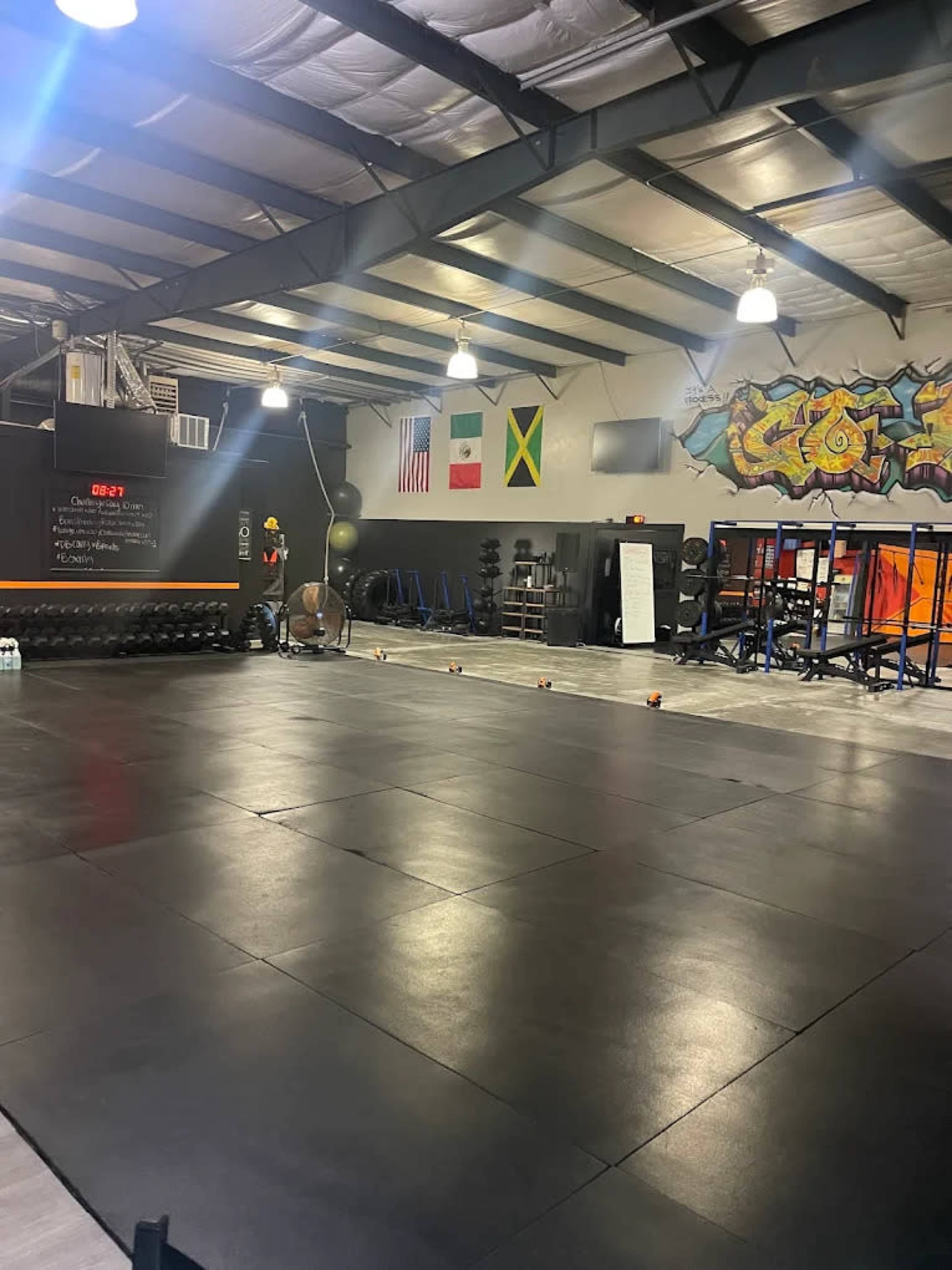 The image shows an empty gym with weightlifting equipment, rubber flooring, and flags of Mexico and Jamaica hanging on the walls, alongside a colorful mural.